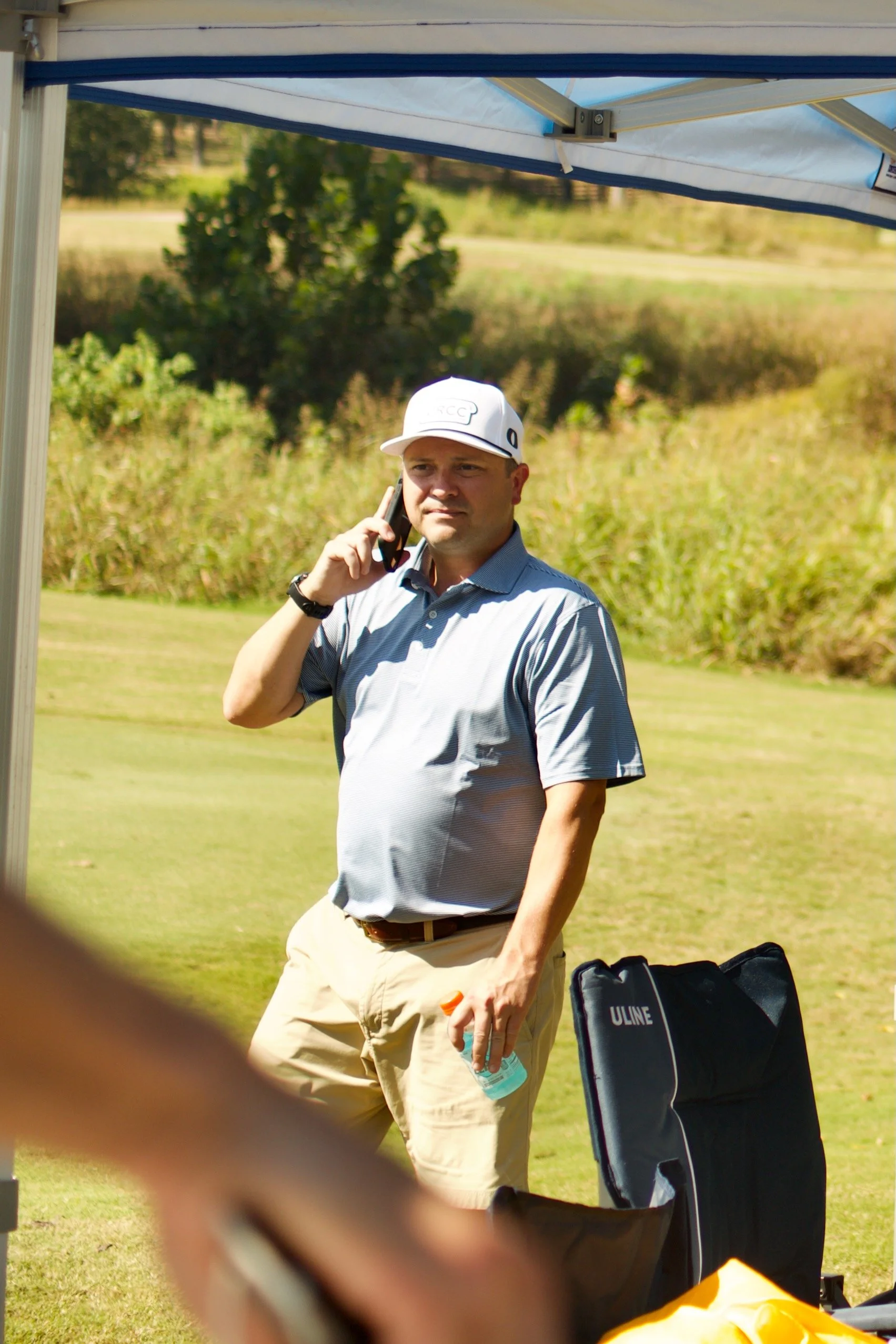 A man with a white cap talking on his cell phone outdoors, standing near a golf bag and equipment on a sunny day.