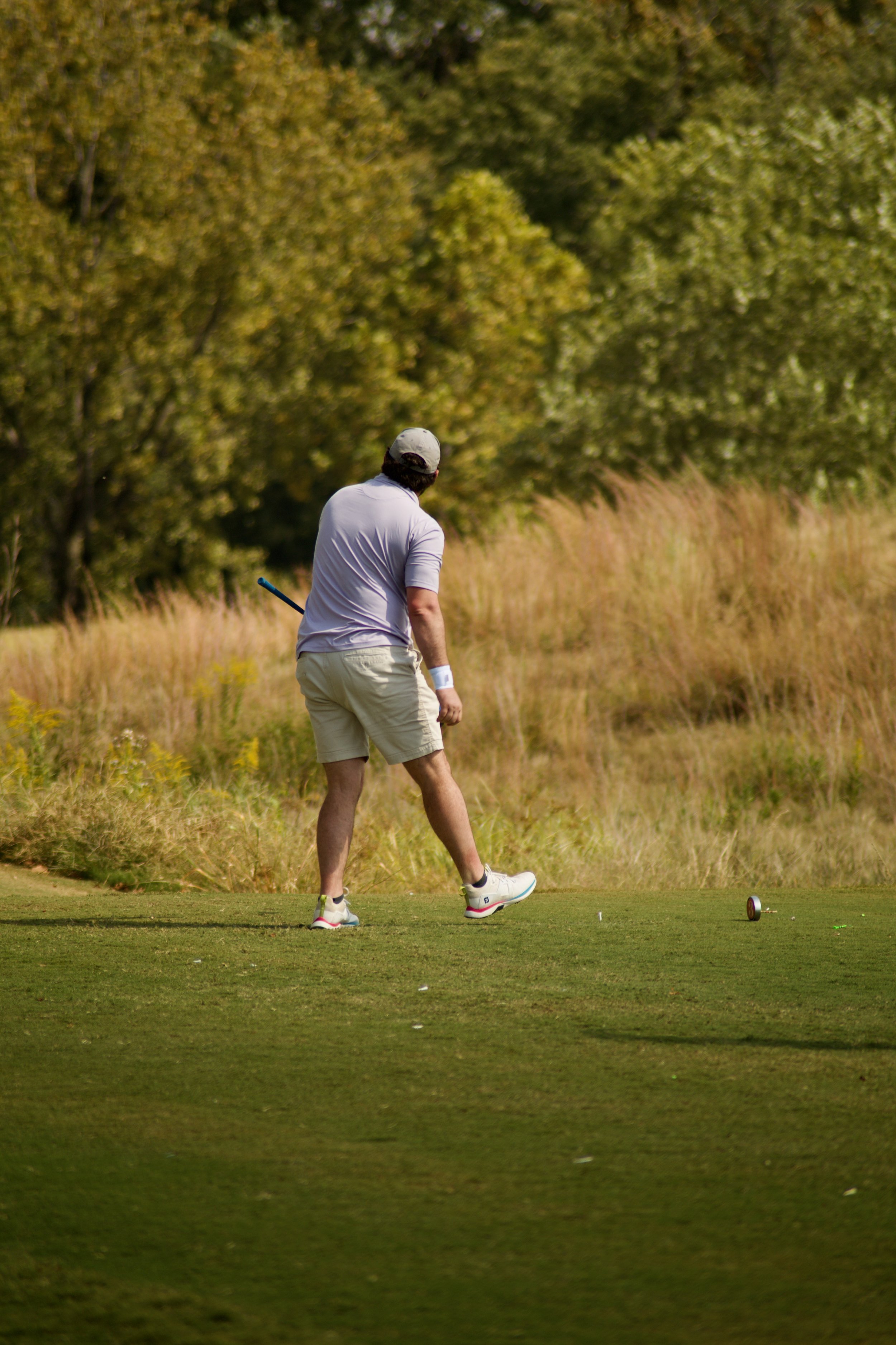 A man wearing a light gray cap, white polo shirt, and beige shorts playing golf on a grassy course with trees in the background.
