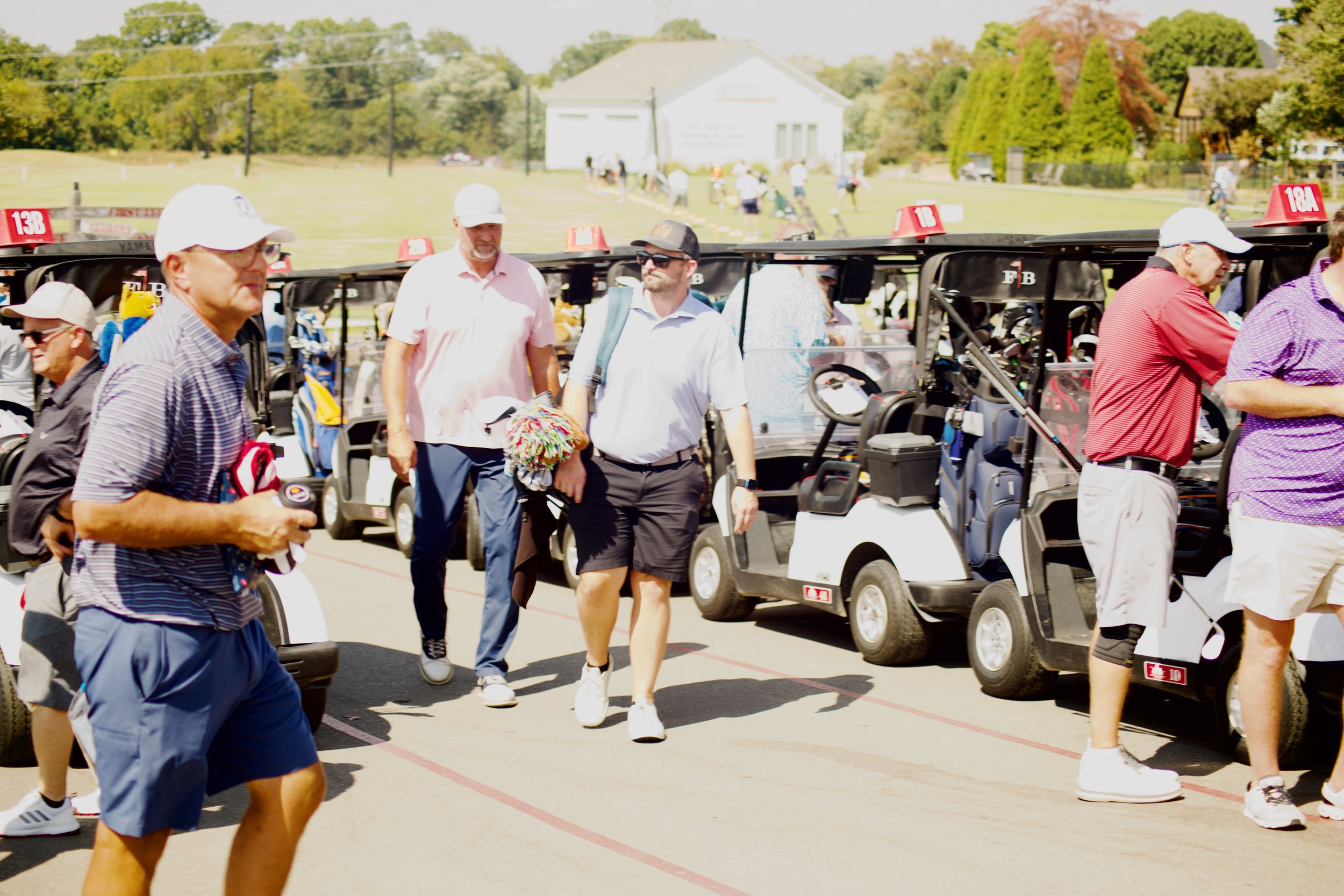 People walking near golf carts on a sunny day at a golf course with a grassy field and small buildings in the background.