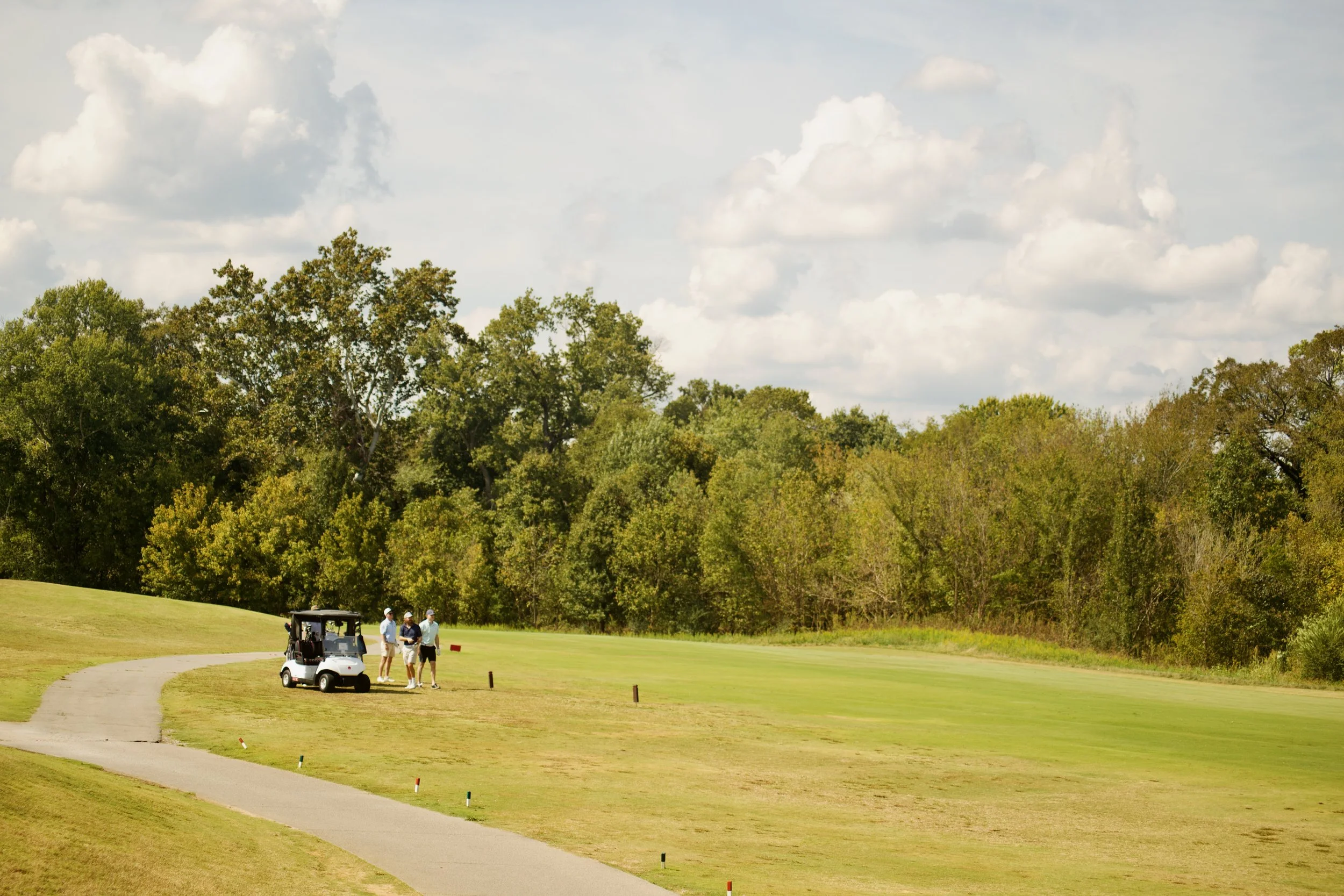 Four people standing near a golf cart on a golf course, with a row of trees and a partly cloudy sky in the background.