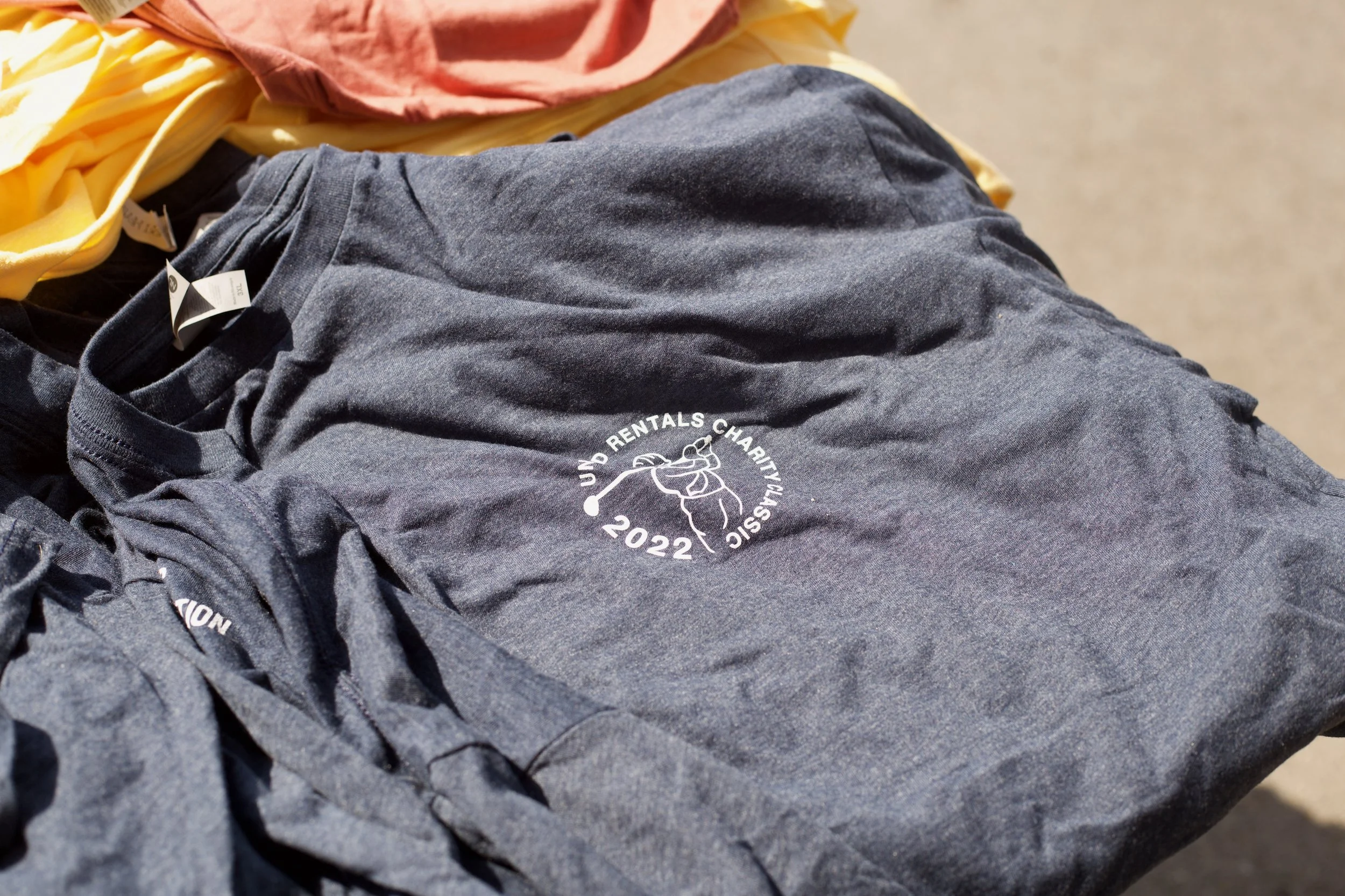 A person wearing a gray T-shirt with a white logo that reads 'URBAN RENTALS CHARITY VALS 2022' and a white outline of a golfer. The person is lying on a sandy surface with yellow and pink clothing nearby.