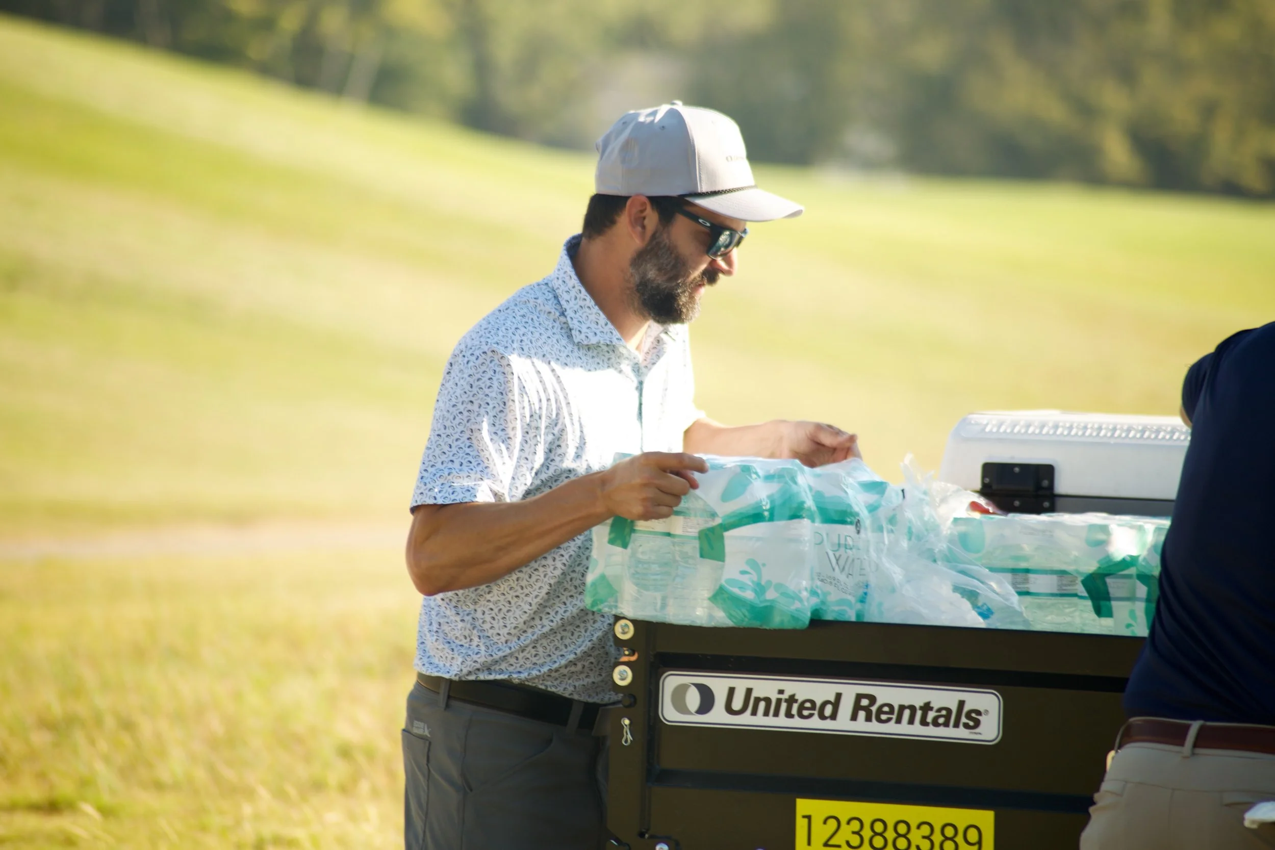 A man with a beard, wearing sunglasses, a white cap, and a patterned shirt, standing outdoors on a sunny day. He is looking at a cooler with a United Rentals logo, surrounded by greenery and bright sunlight.