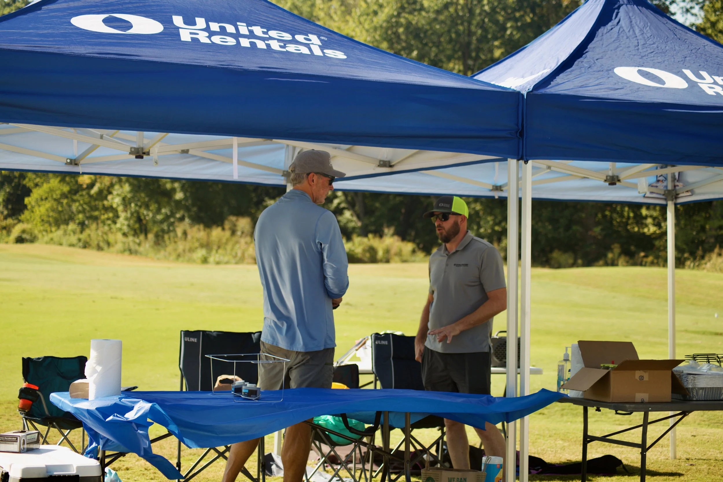 Two men standing and talking under a blue United Rentals canopy tent outdoors on a sunny day with trees in the background. Various equipment and supplies are on the table and around them.