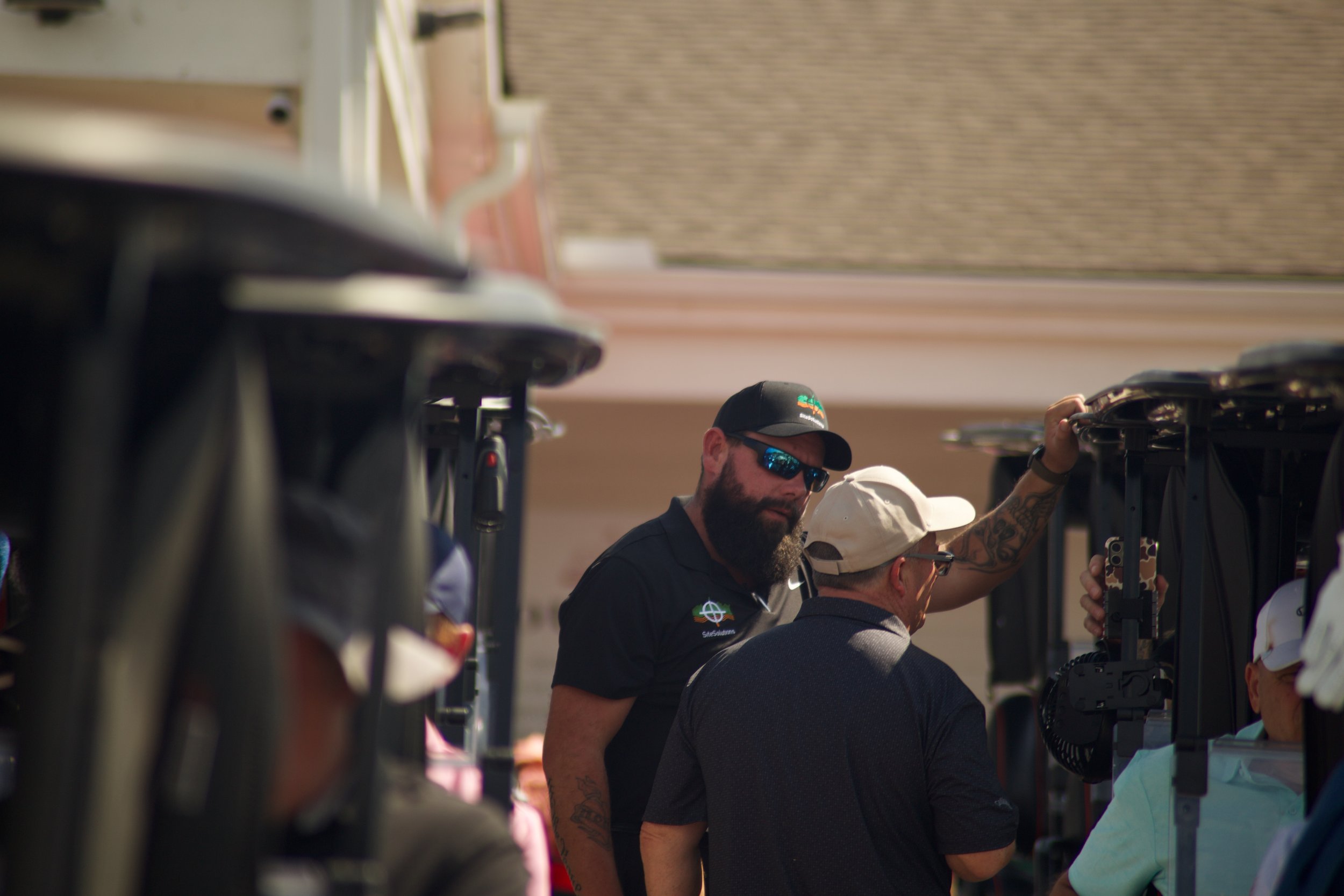 People are working on golf carts at an outdoor event, some are wearing sunglasses, caps, and casual clothing.