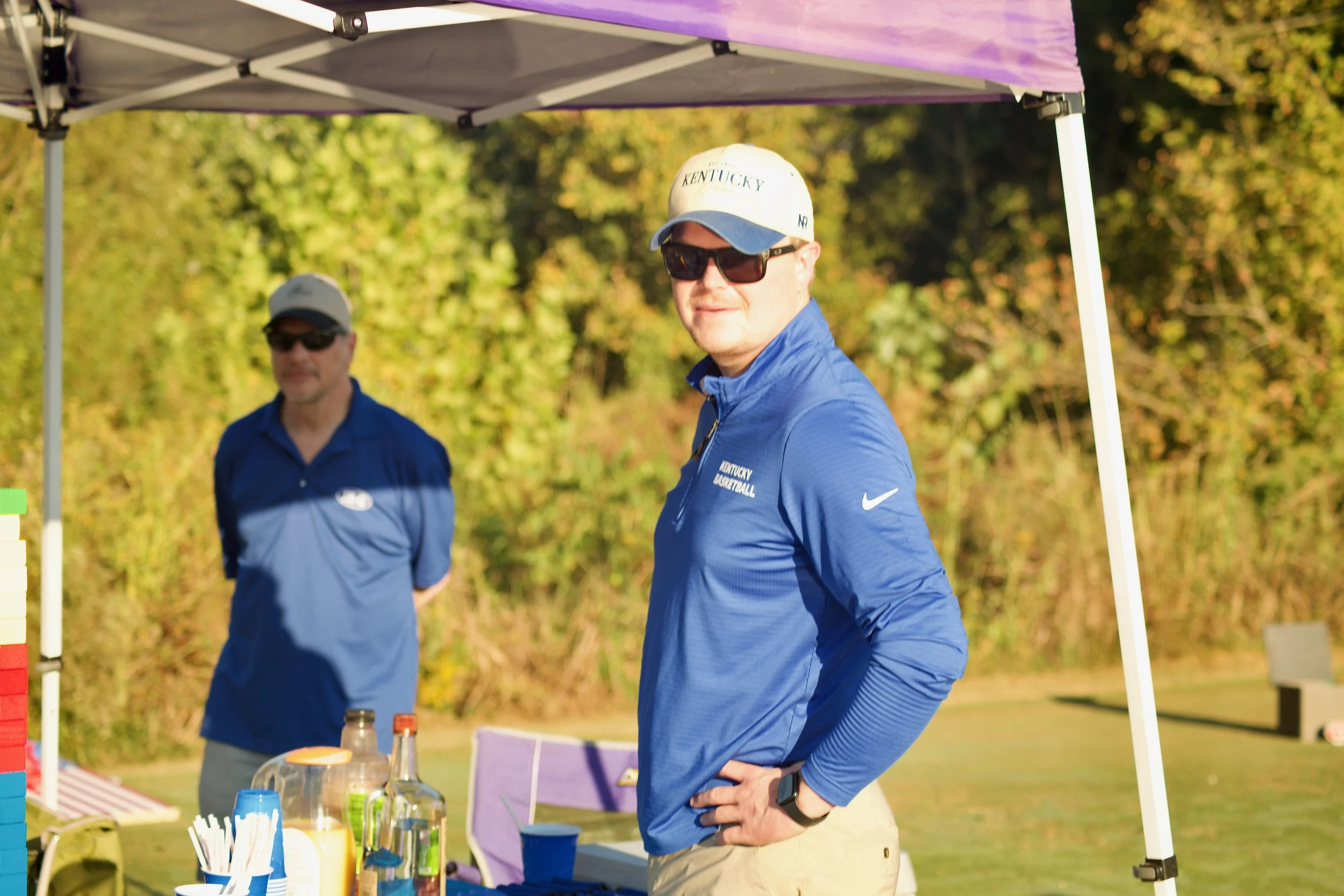 Two men wearing Kentucky Wildcats shirts and hats, standing under a canopy at an outdoor event on a sunny day with trees in the background.