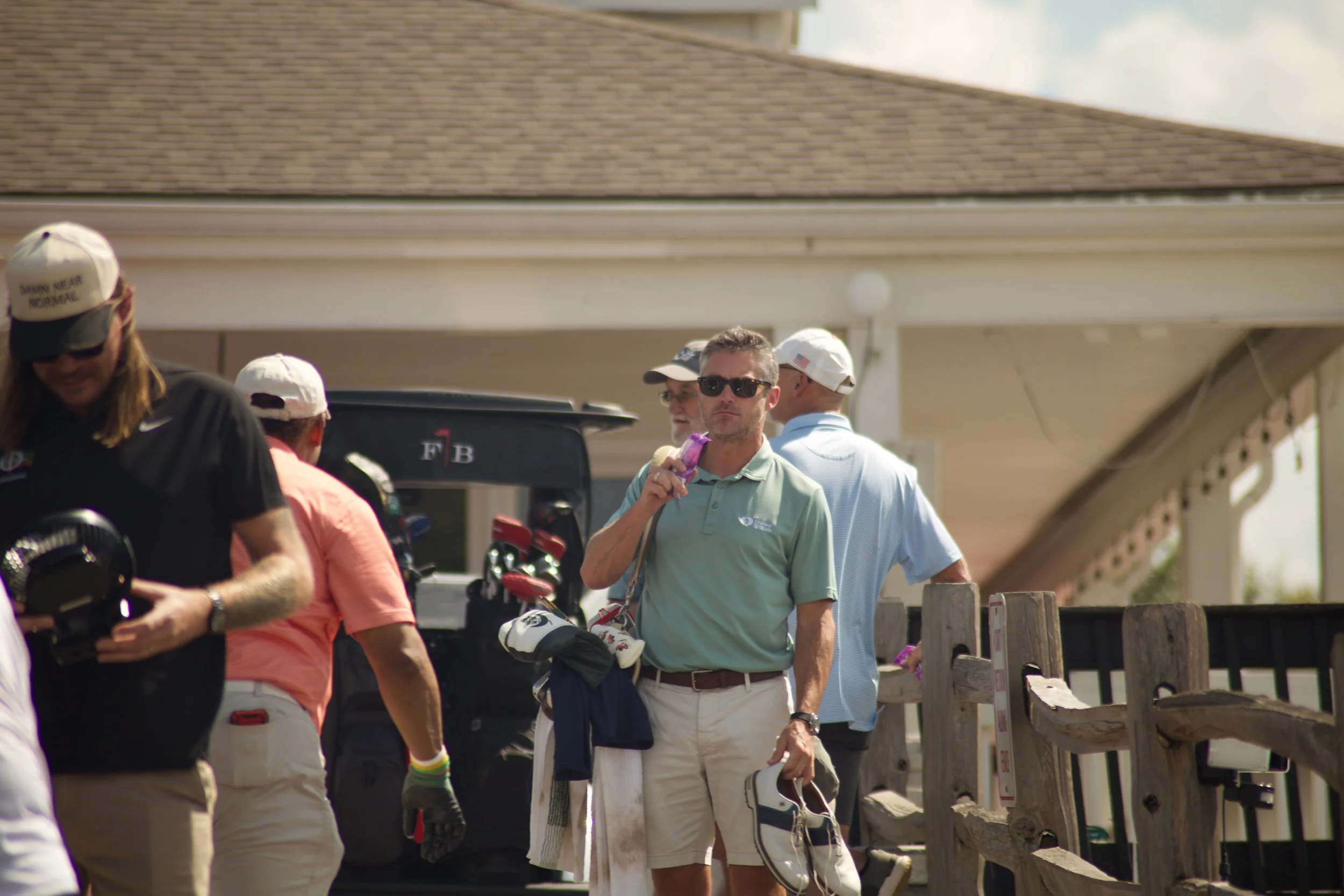 Group of people at a golf course, some holding golf clubs and wearing golf attire, standing near a wooden fence.