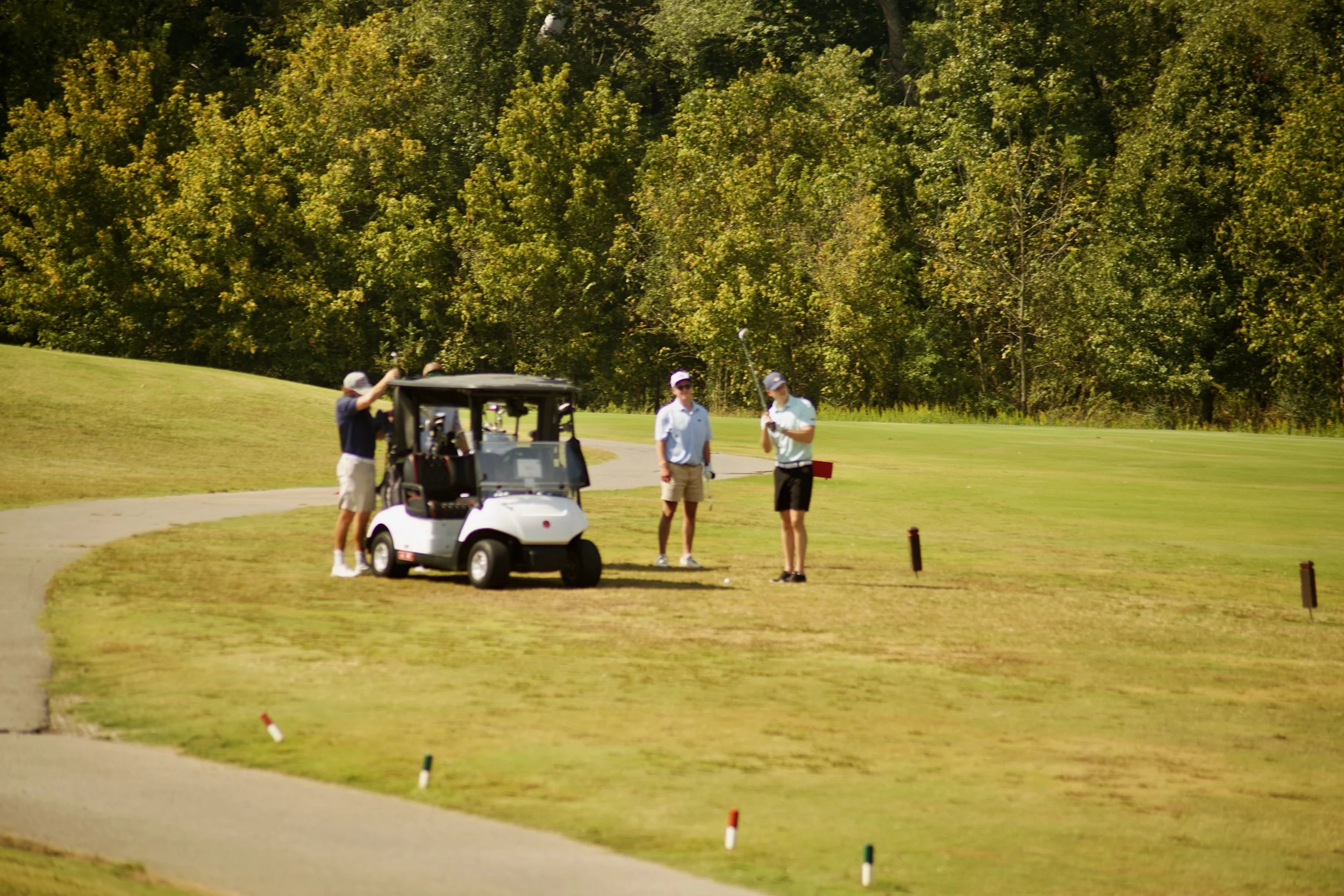 Three people on a golf course near a golf cart, two of them with golf clubs, with large trees in the background.