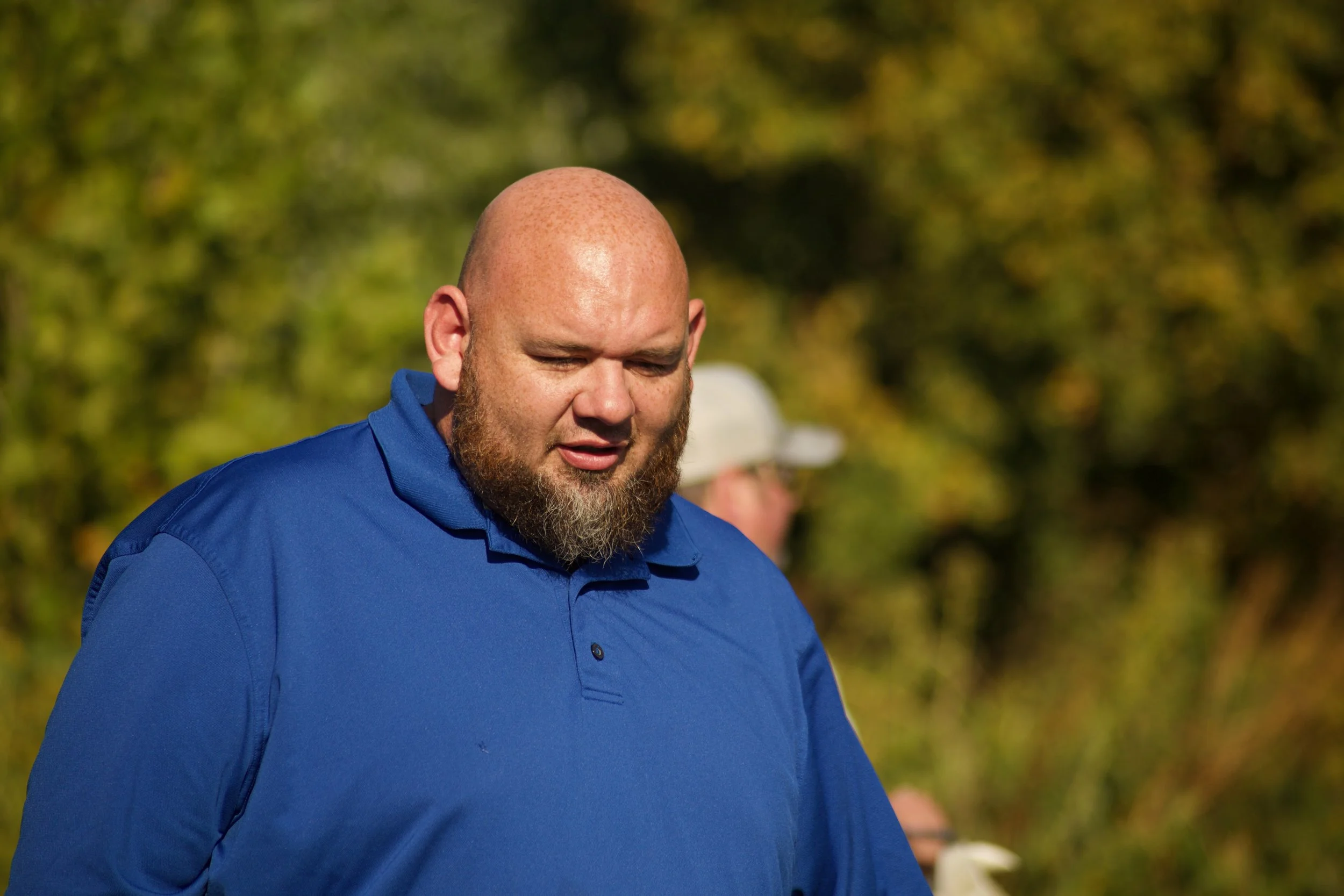 A bald man with a beard wearing a blue collared shirt outdoors with a background of green trees.
