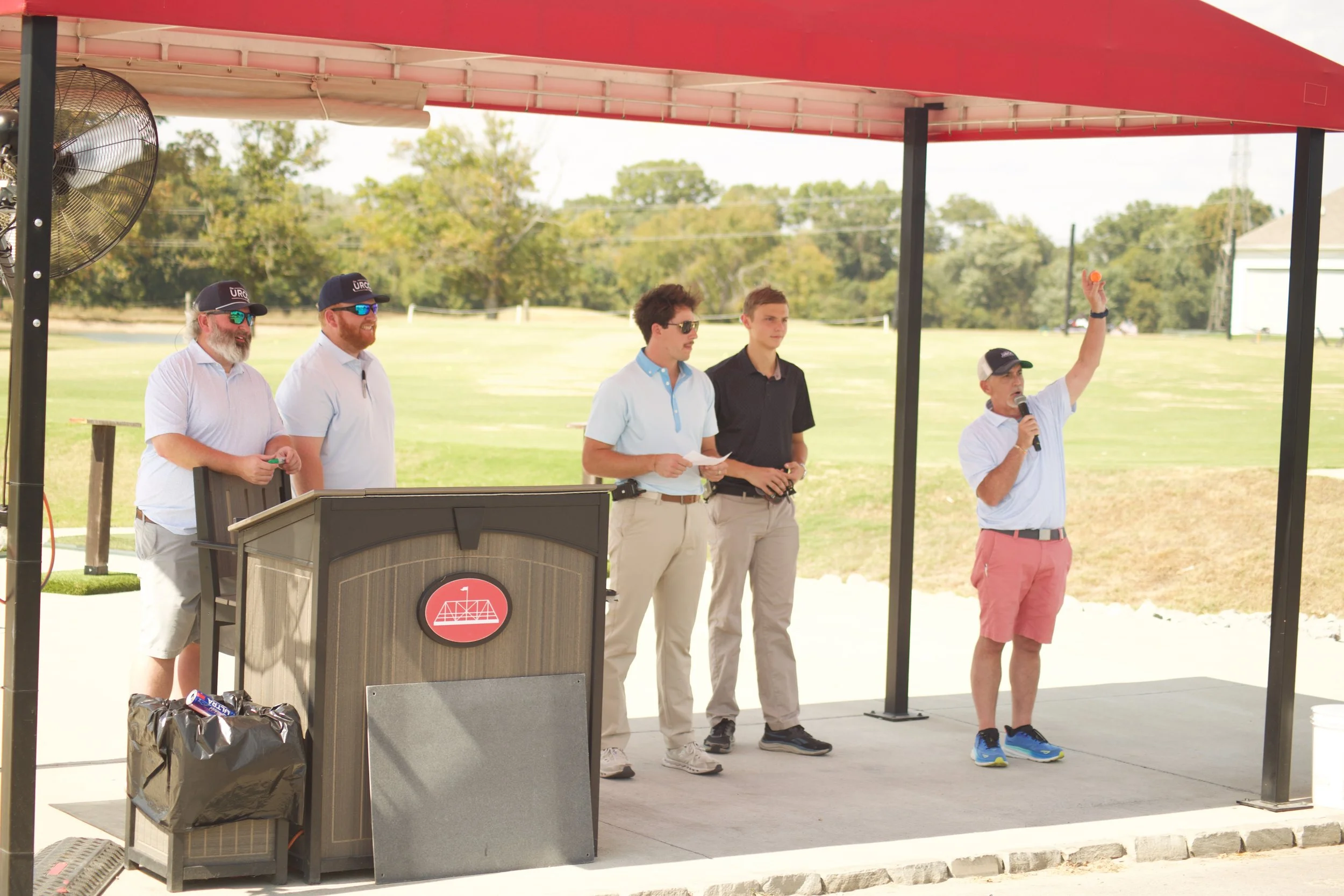 A group of five men standing and speaking at an outdoor event under a canopy, with four men wearing baseball caps and one man holding a microphone and a small orange ball, in a park or open field.