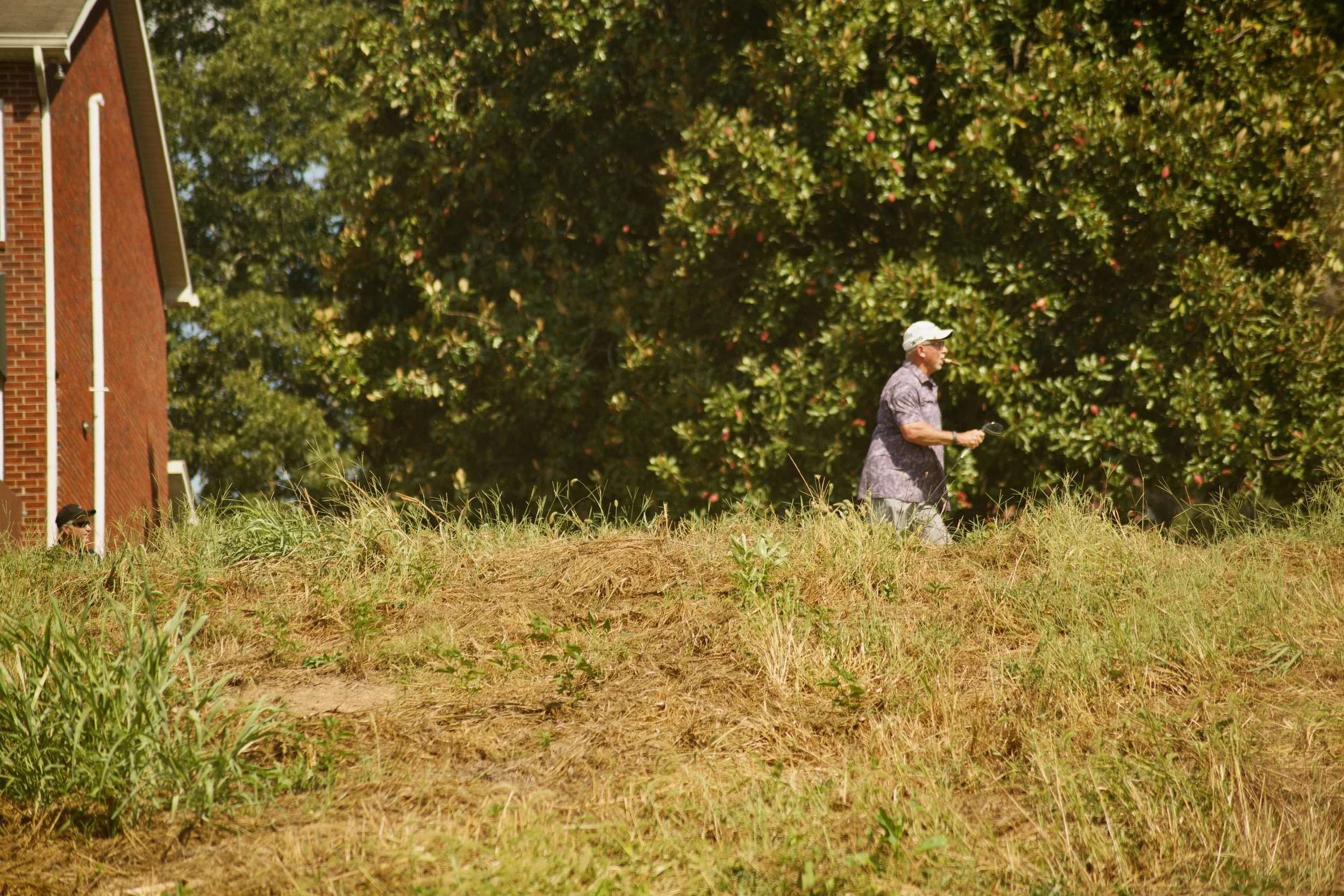 A man wearing a hat and sunglasses walking outdoors in a grassy area with trees and a brick building in the background.