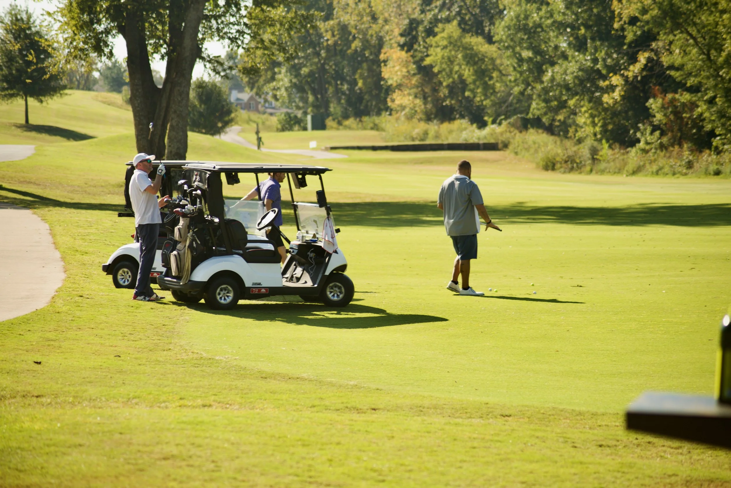 Two men playing golf on a sunny day, one standing near a golf cart and the other preparing to hit the golf ball, with trees and a golf course in the background.