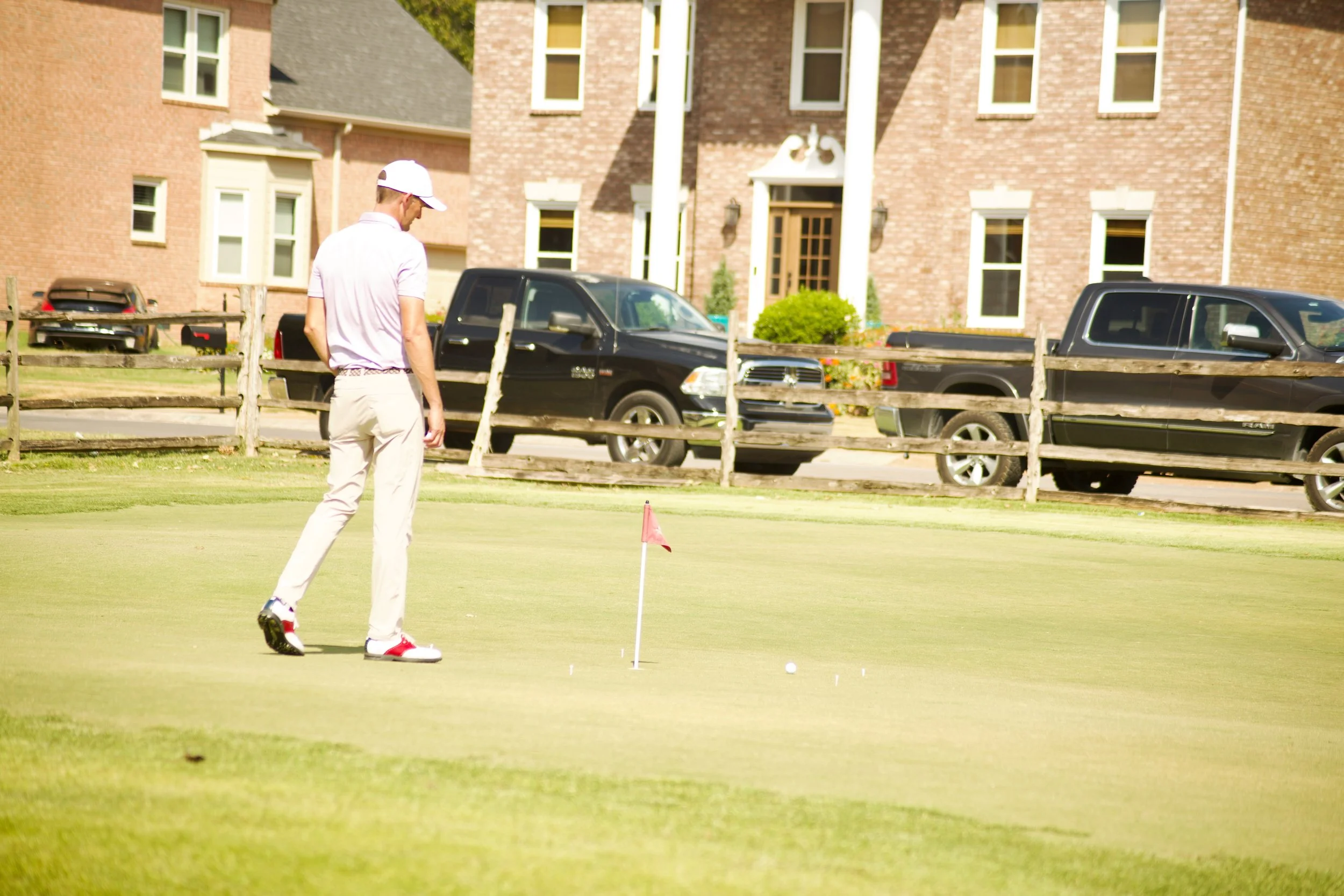 A man playing golf on a green, preparing to putt, with a flagstick and golf ball on the short grass. In the background, there are several parked cars and a multi-story brick residential building.
