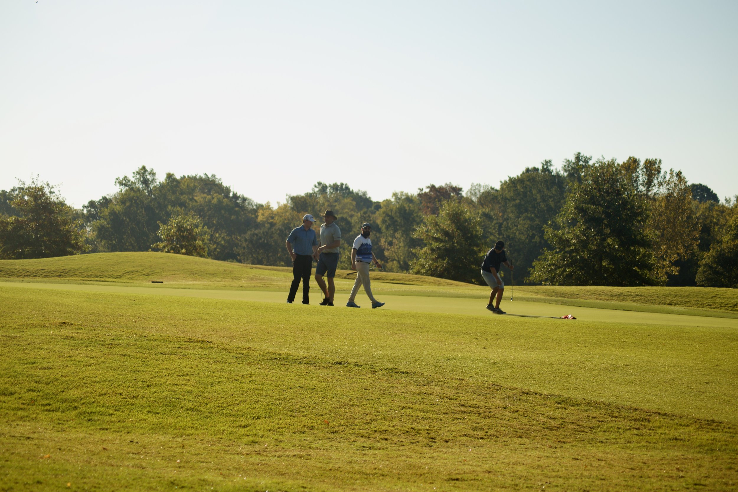 Four men on a golf course, two of them are standing and two are preparing to putt, with trees in the background and a clear sky.