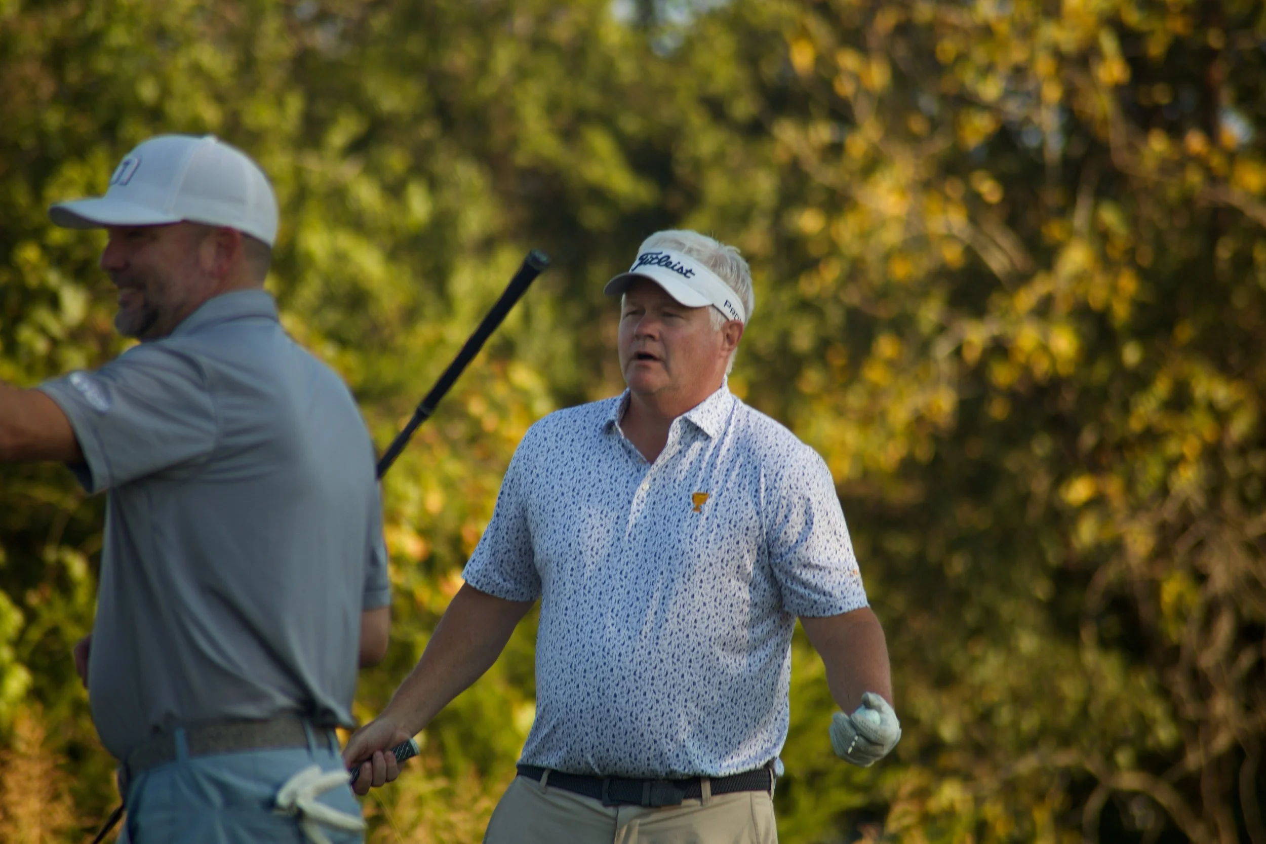 Two men on a golf course, one in a white cap with a Titleist logo and the other with a patterned shirt and a visor, holding hands and walking with golf clubs, surrounded by trees with autumn leaves.