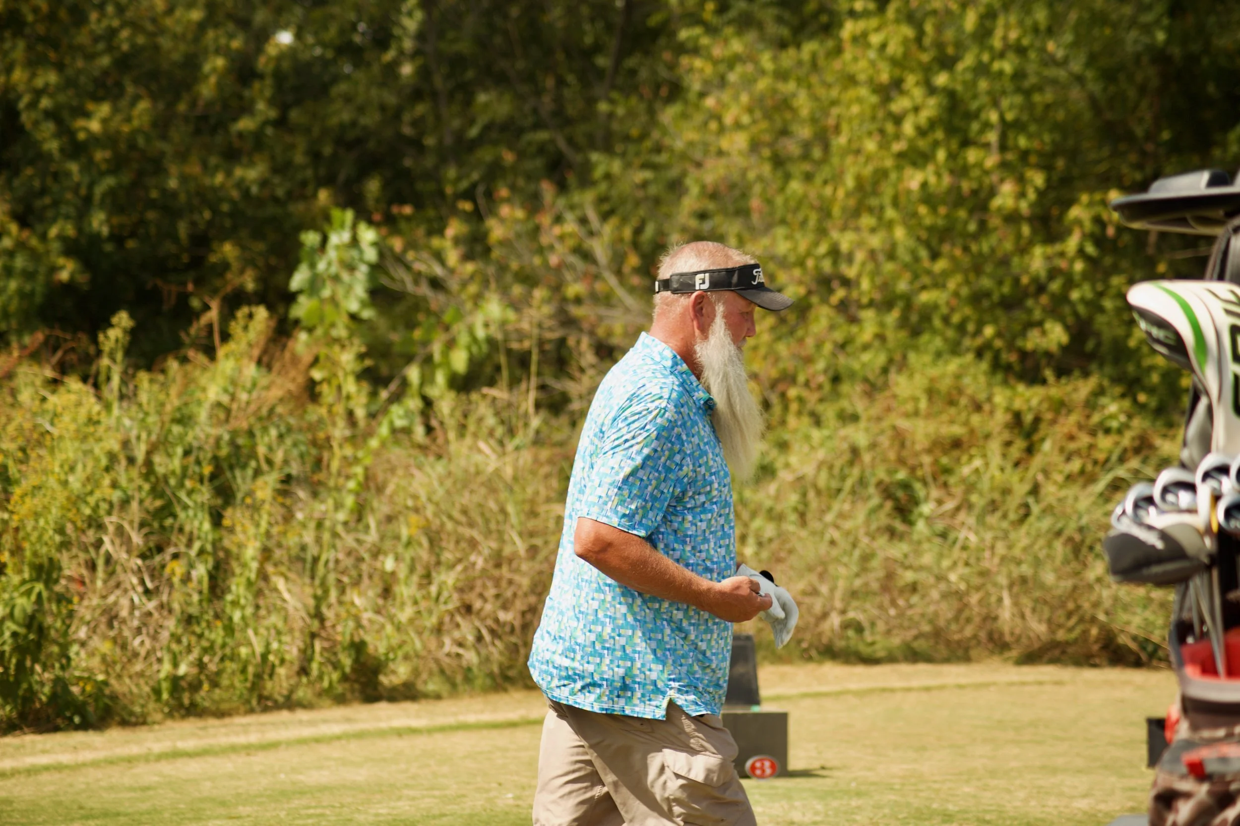 An older man with a long white beard, wearing a black visor and a blue patterned polo shirt, stands outdoors with a golf bag and golf equipment nearby, surrounded by trees and greenery.