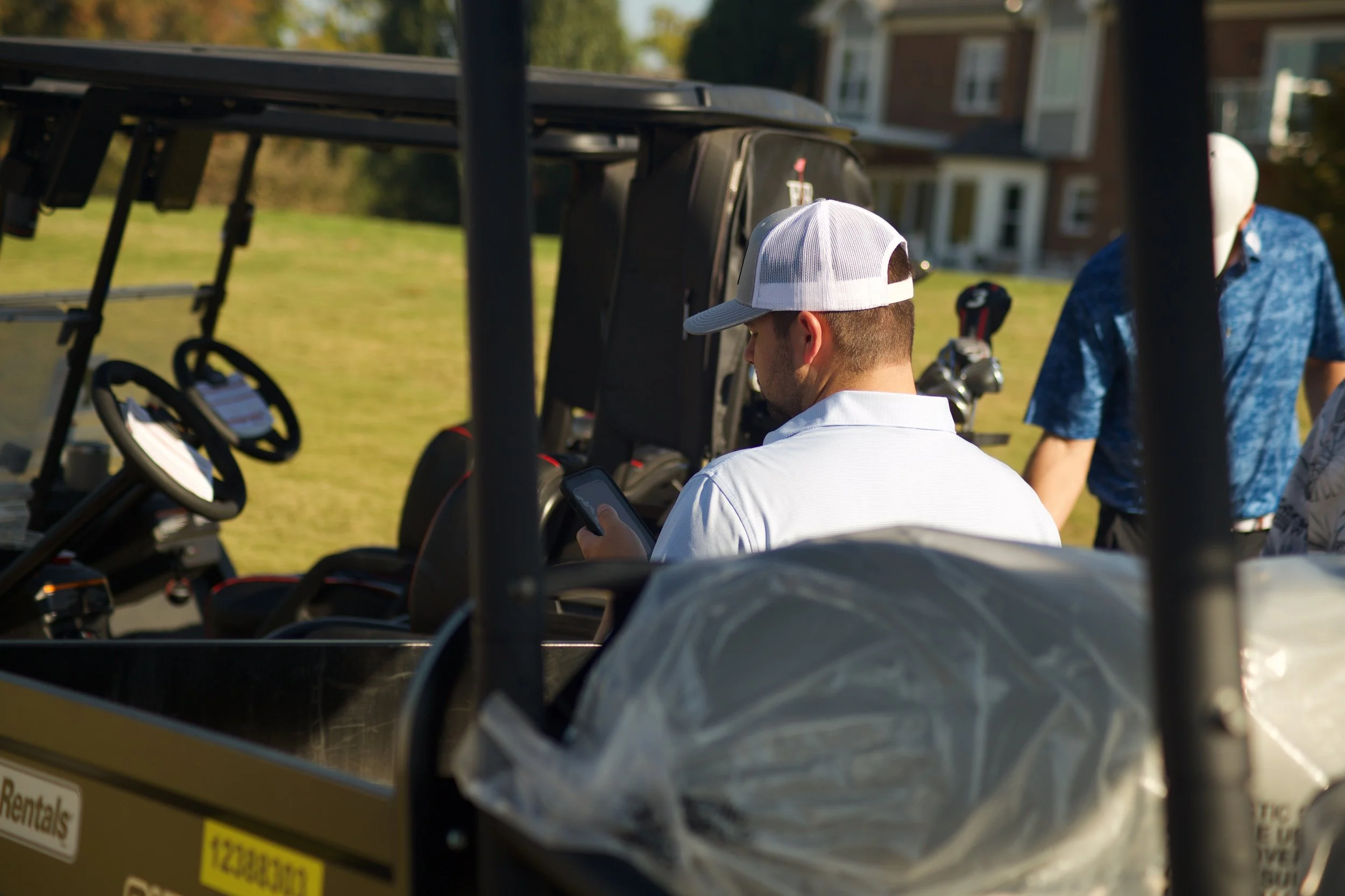 A man wearing a white baseball cap and white shirt looking at his phone on a golf cart. There are other people nearby, and the background shows a grassy area with trees and a brick building.