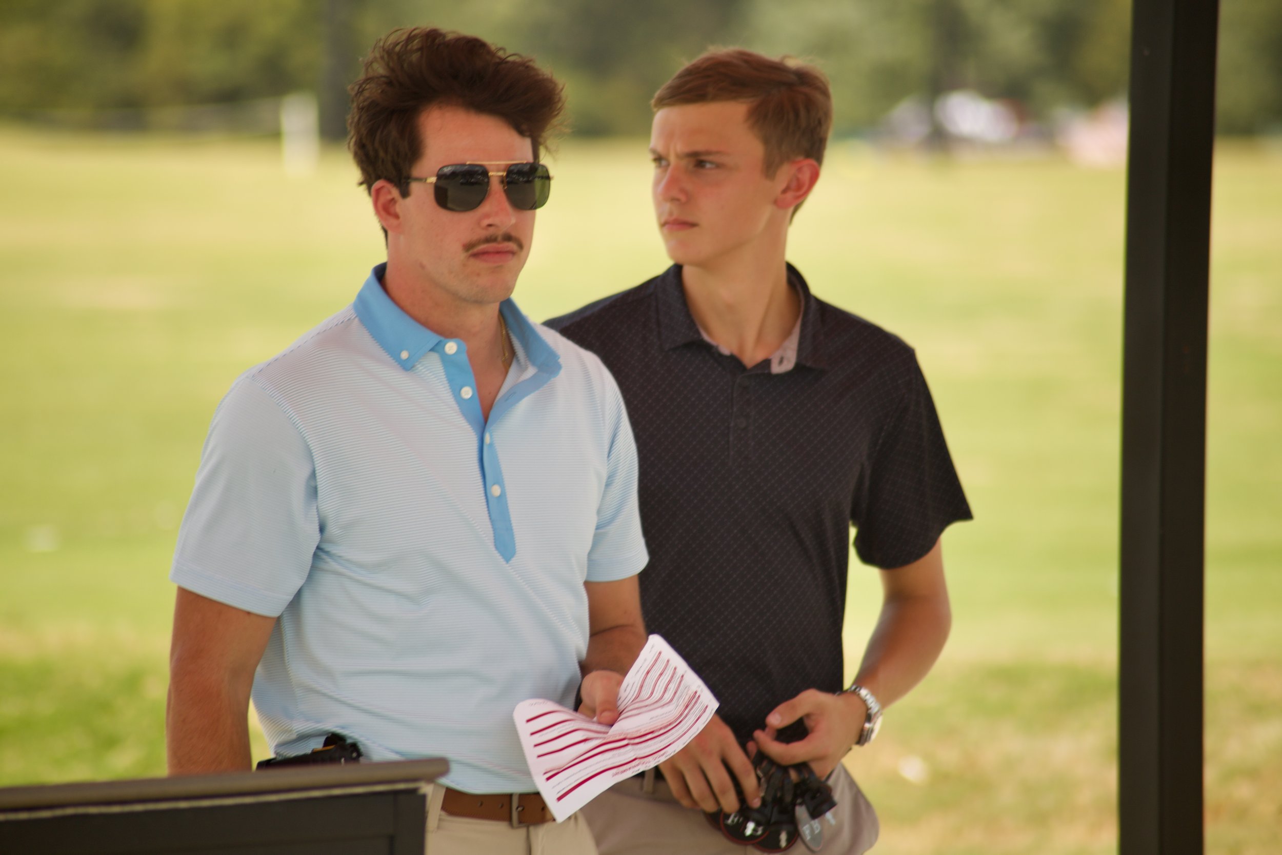 Two young men standing outdoors in a park, one wearing sunglasses and a light blue polo shirt holding a striped paper, the other in a black shirt holding a camera, with a grassy field and trees in the background.