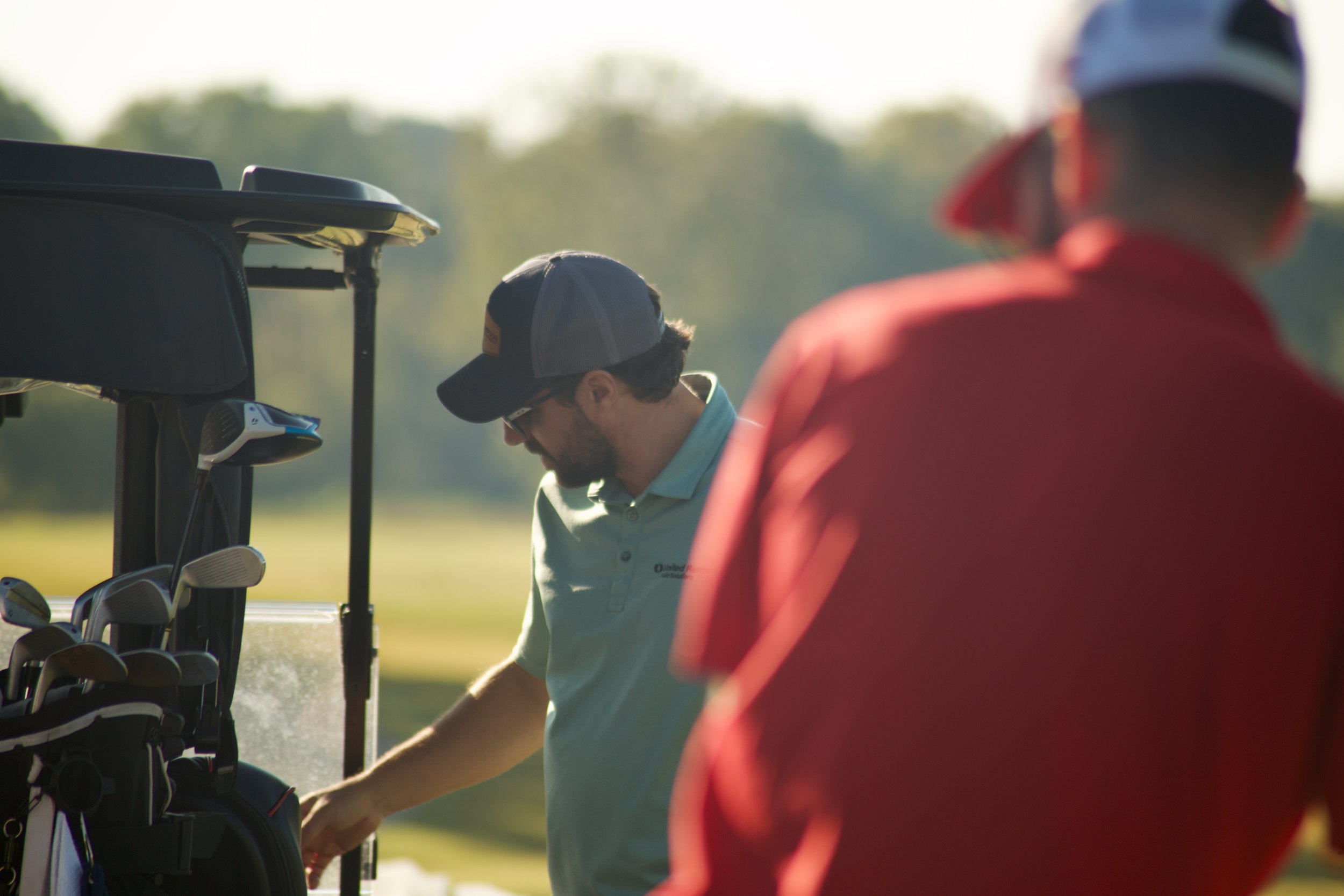 A man at a golf course standing near a golf bag with clubs, talking with a woman wearing a red jacket and helmet.