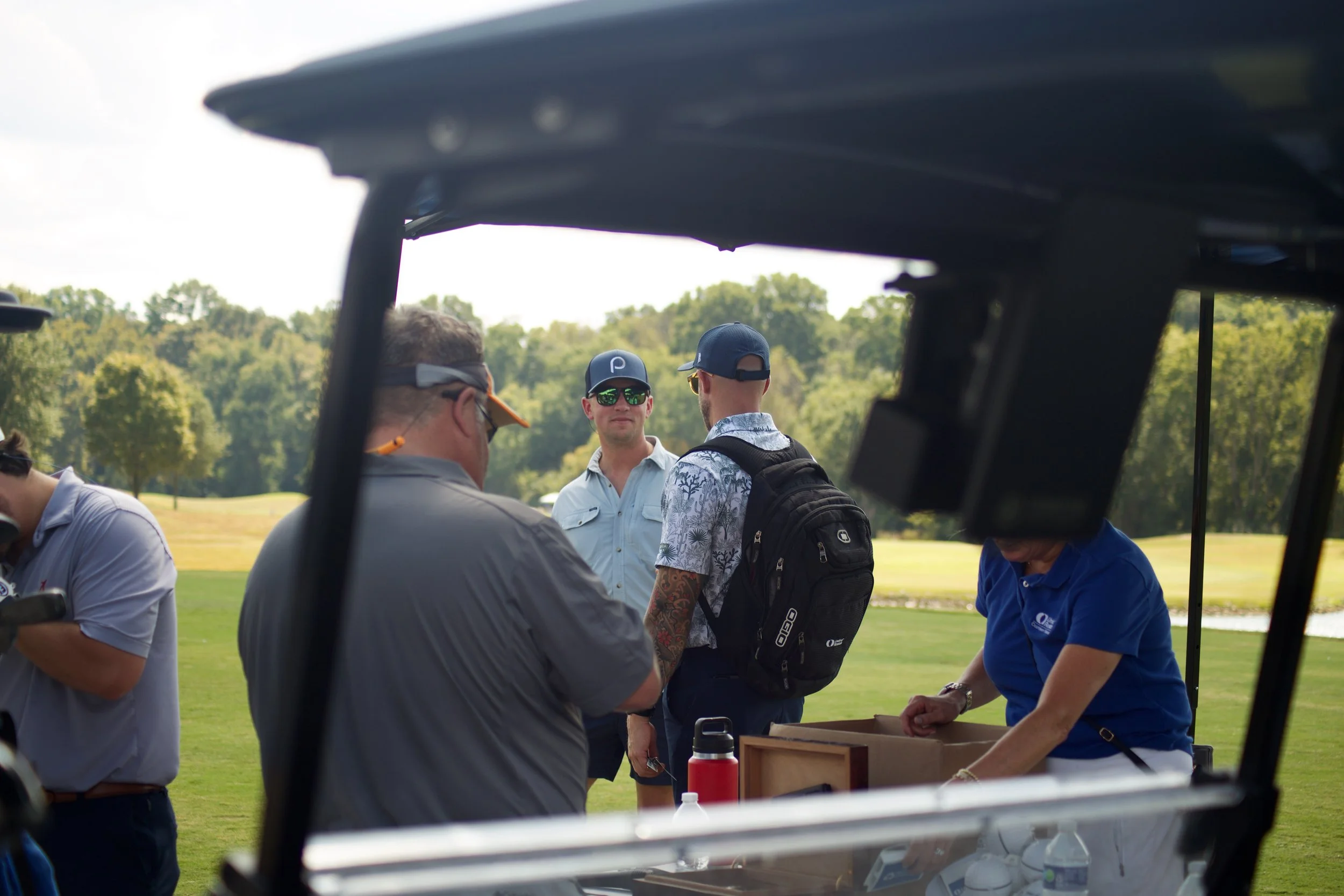 Group of men gathered outdoors on a golf course, engaged in conversation, with trees and a partly cloudy sky in the background.