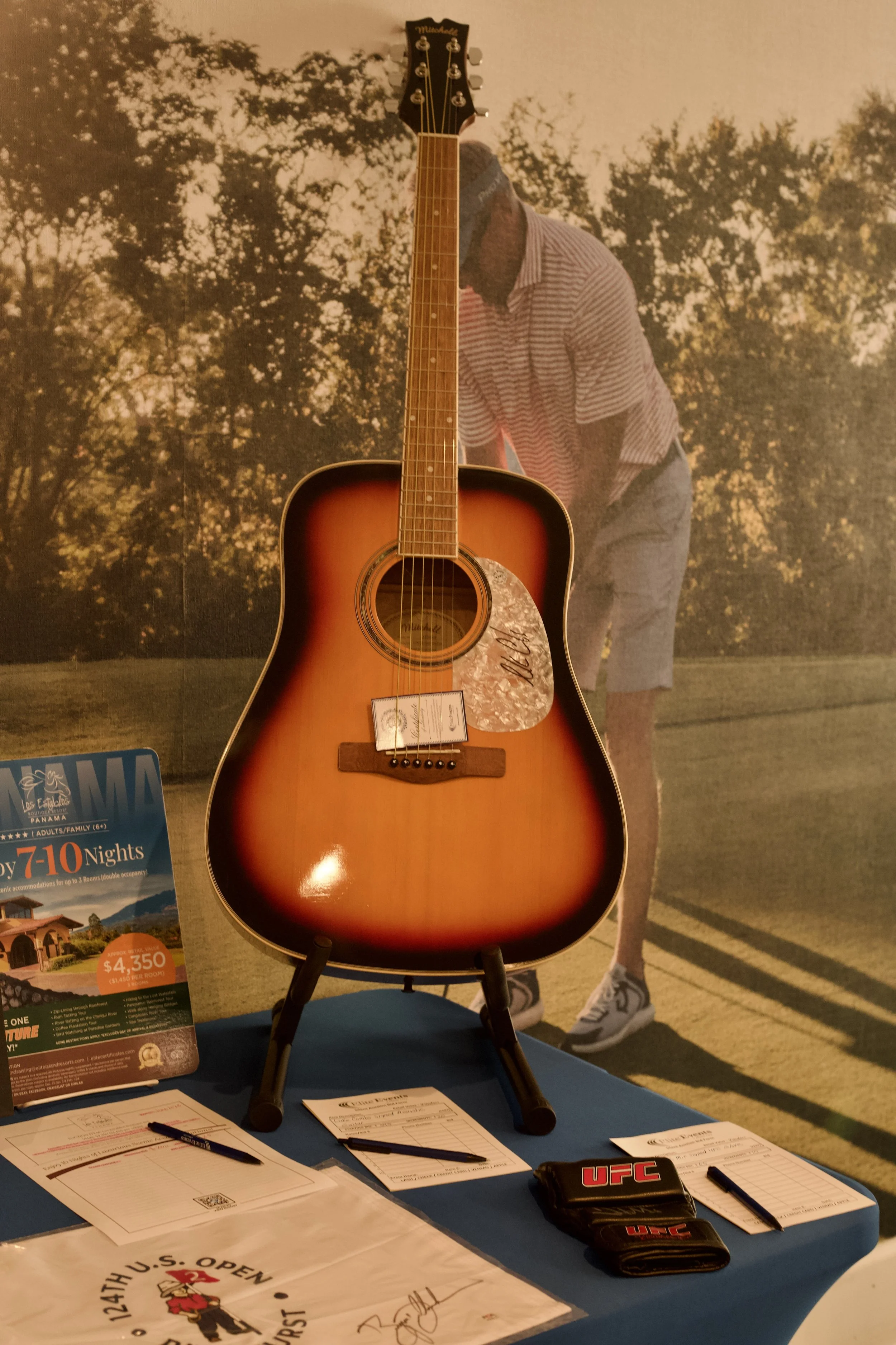 An acoustic guitar displayed on a table with paperwork, pens, UFC branded items, and a poster advertising a travel deal in front of a large sepia-toned image of a person in a striped shirt and shorts outdoors.