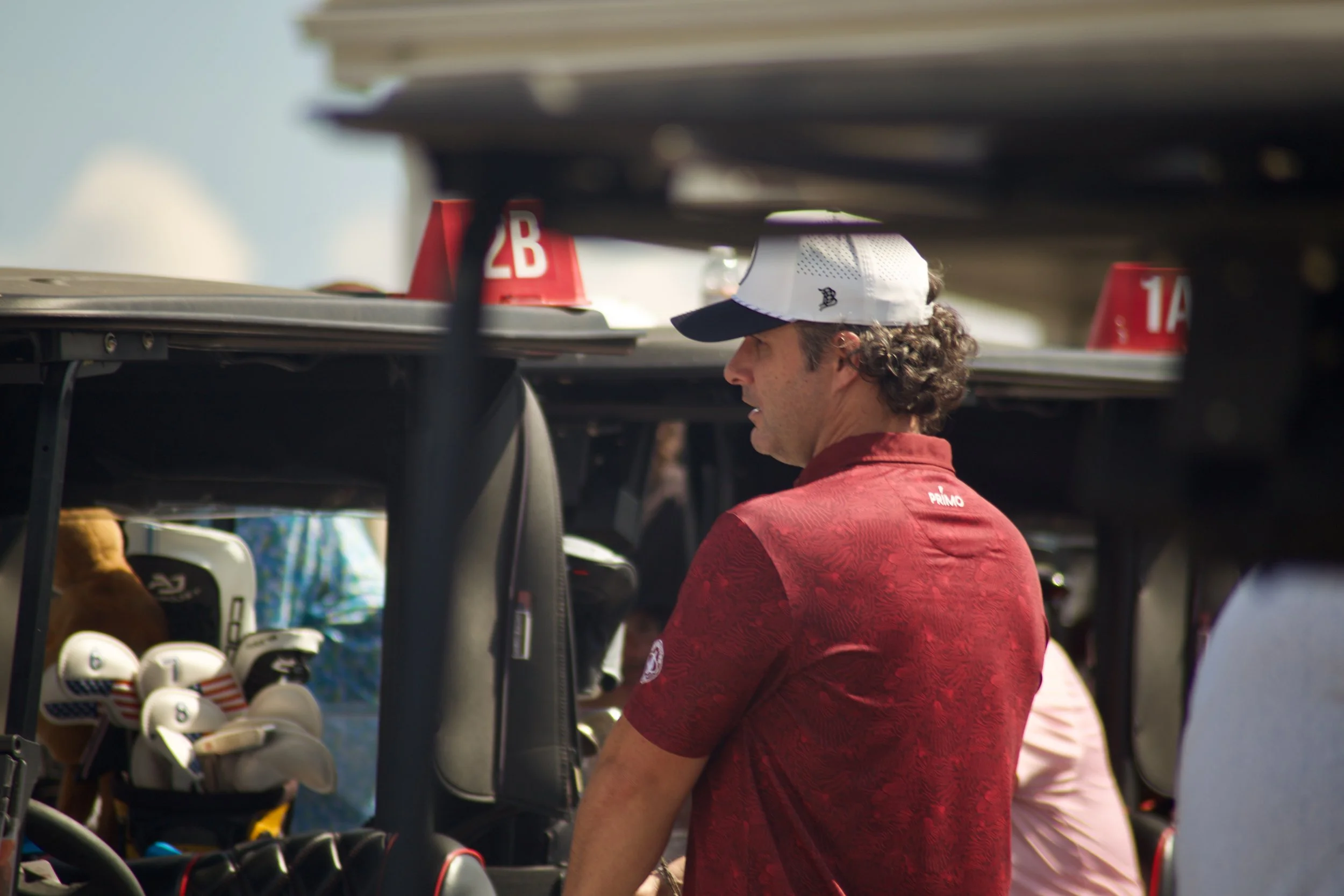 A man with curly hair wearing a white cap and red shirt looking at golf clubs in a golf equipment display at an outdoor event.