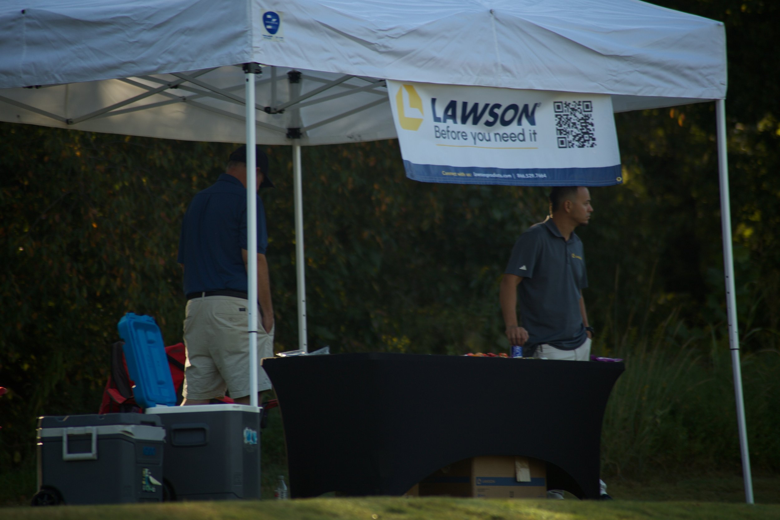 A white canopy tent with a Lawson sign that says 'Before you need it' and a QR code. Two men are standing underneath the tent, with one facing left and the other facing right, surrounded by various boxes and equipment outdoors.
