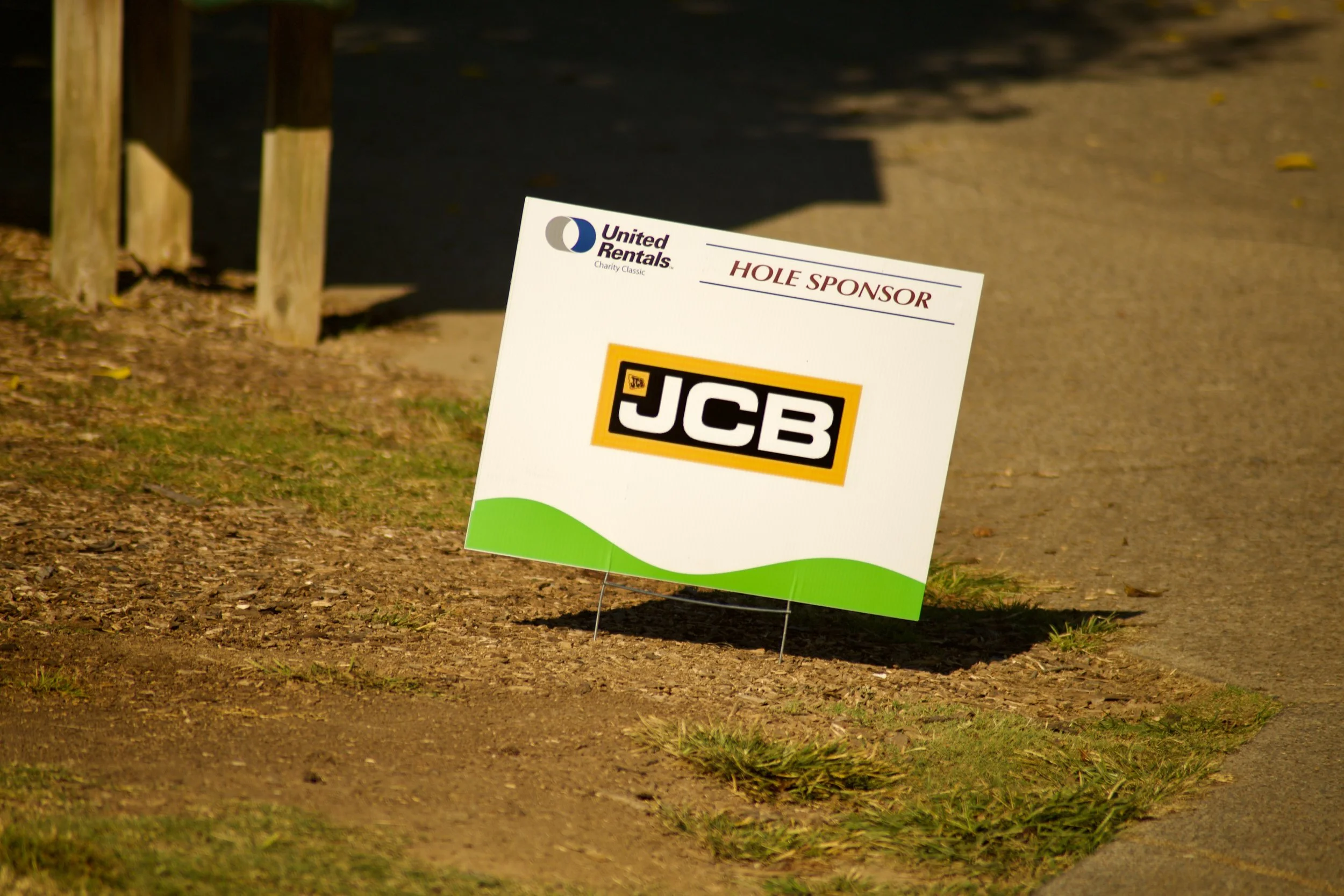 A signboard on a patch of ground with grass, featuring a logo for United Rentals, the words 'HOLE SPONSOR', and a JCB logo below.