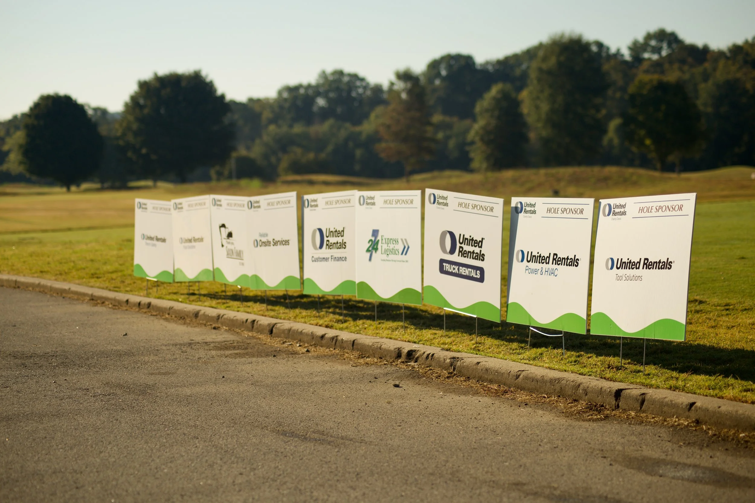 A row of outdoor signs with company logos and sponsorship information lining a grassy area beside a paved road, with trees in the background.