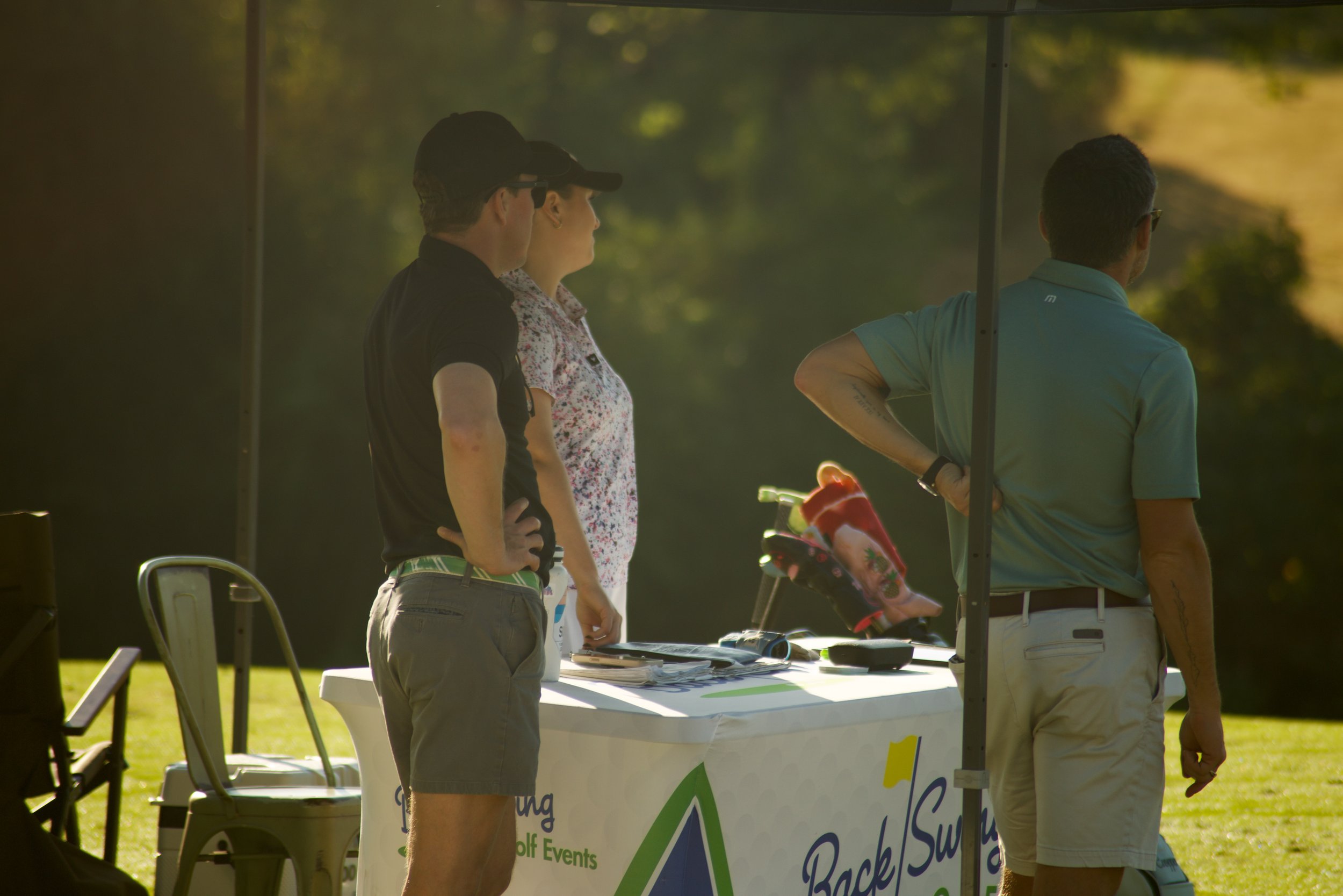People standing around a registration desk at a golf event outdoors in sunlight.