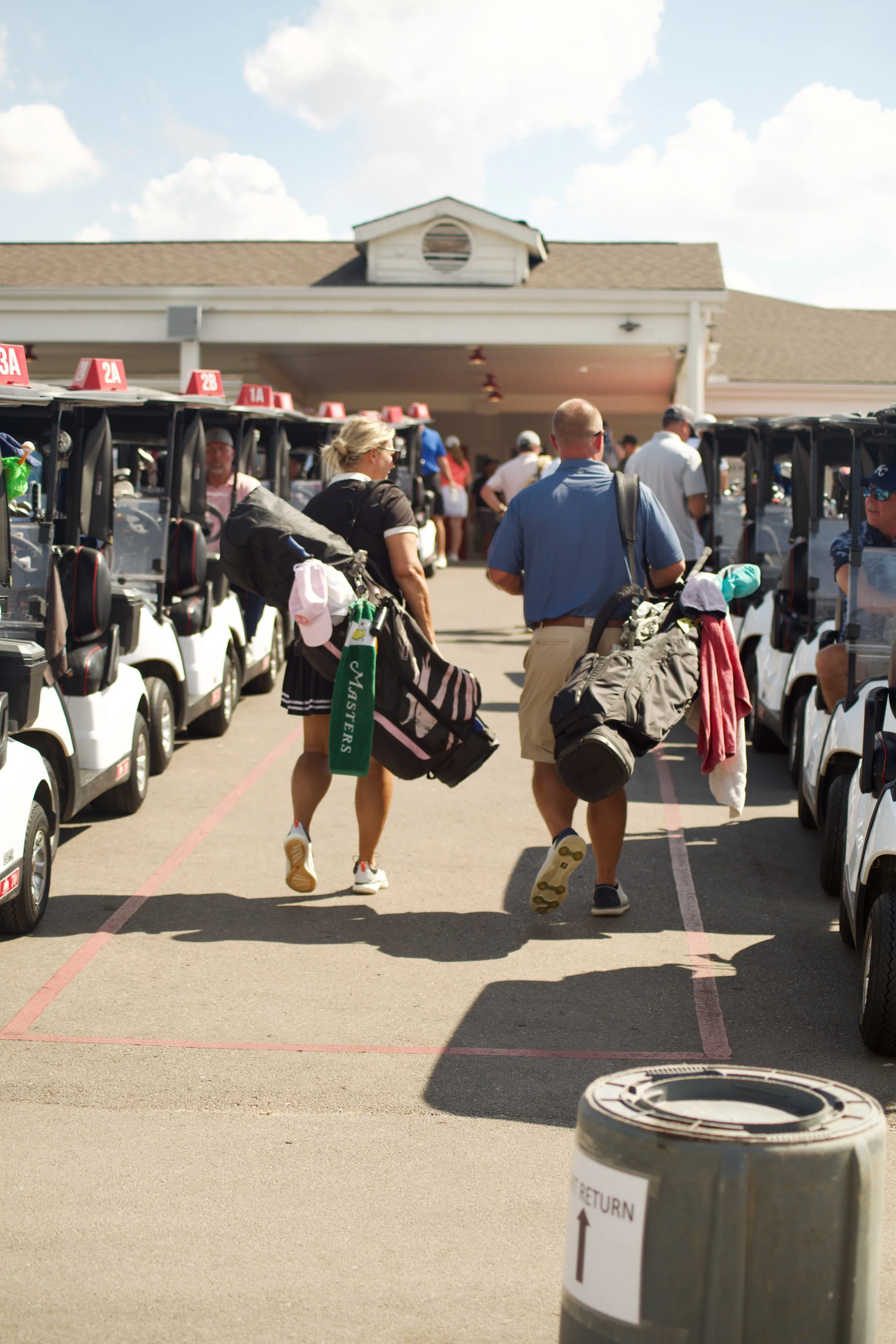 A group of people walking with golf bags toward a building at a golf course, with golf carts lined up on either side.