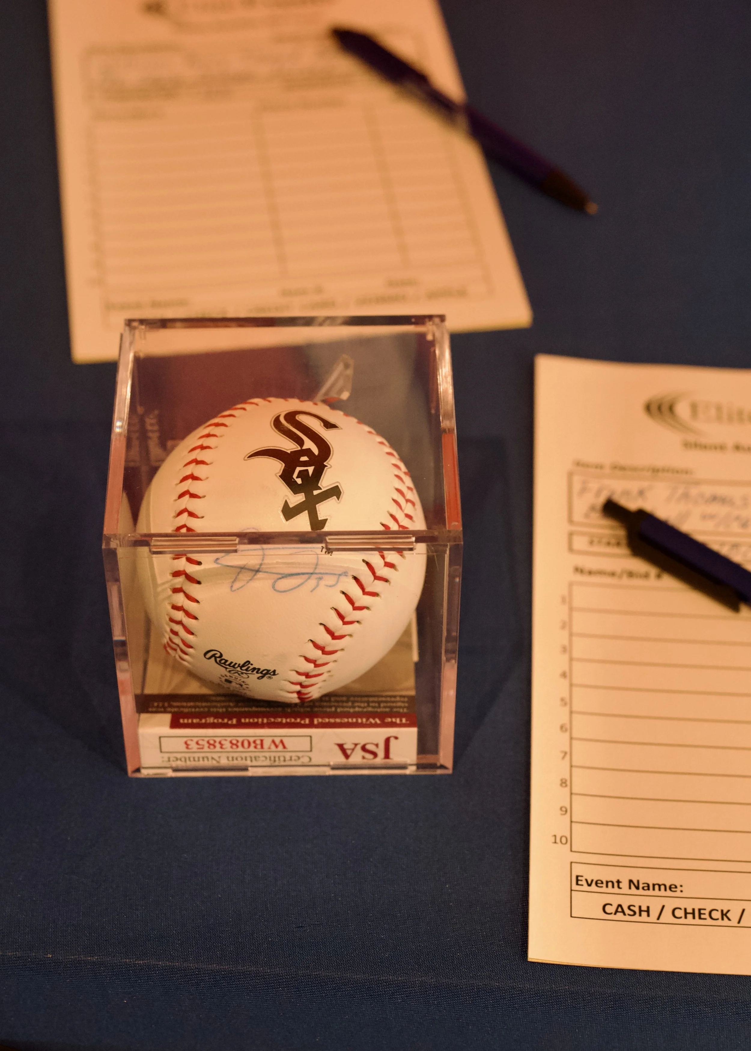 A signed baseball in a clear display case with a black logo. The baseball is placed on a table with event forms and pens nearby.