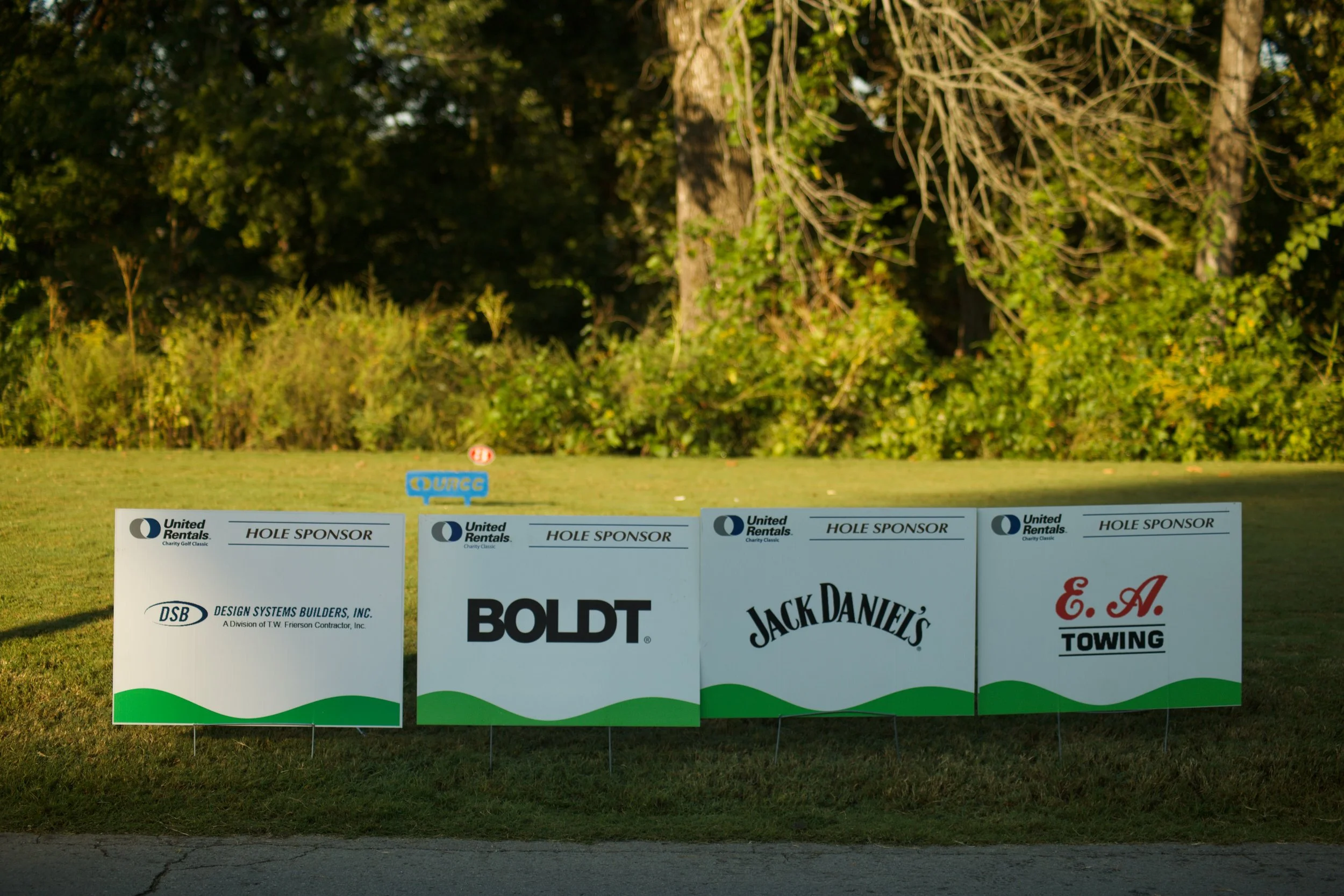 Banners for a charity golf event featuring sponsor logos, set outdoors with trees in the background.