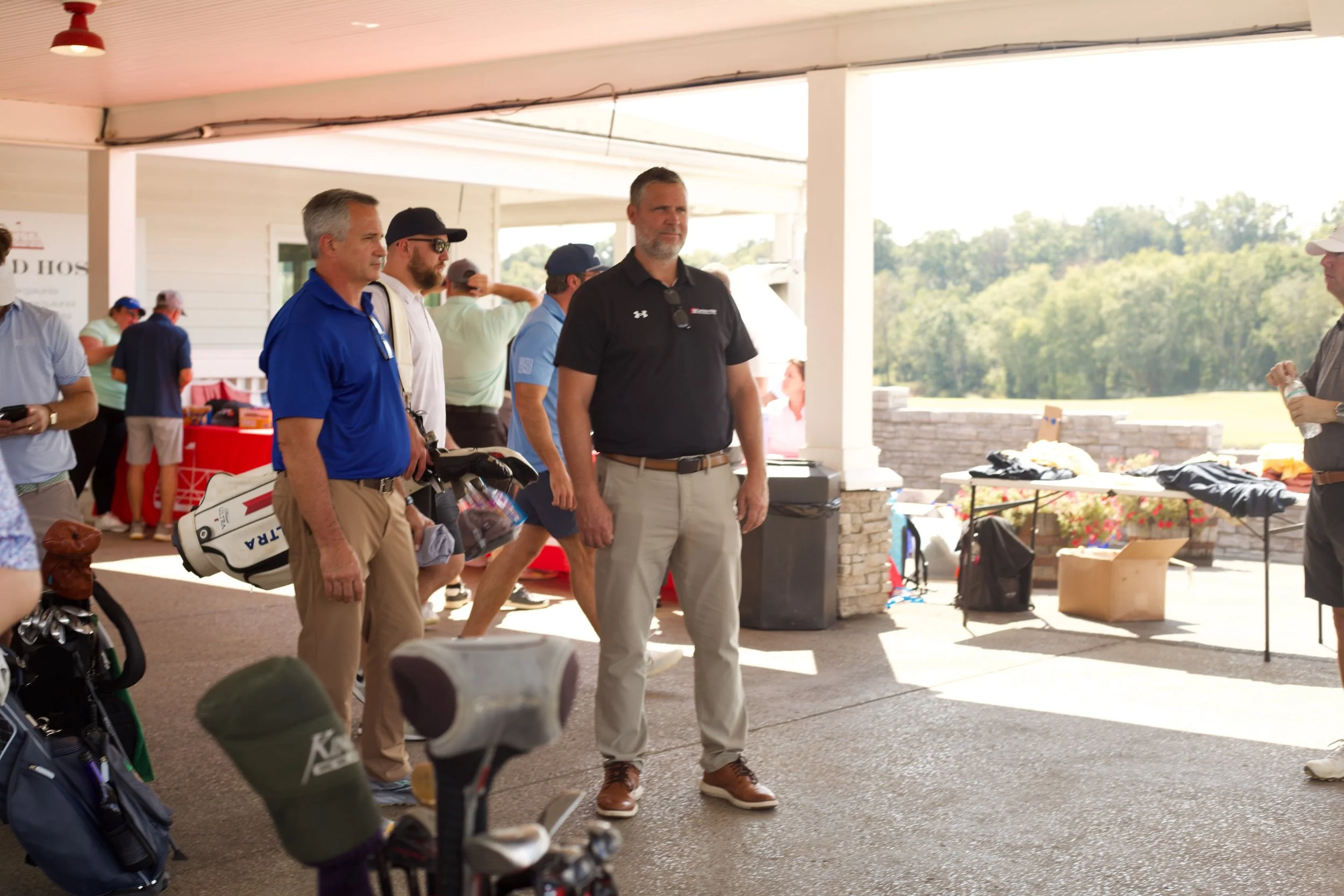 A group of men standing outdoors on a patio or deck area near golf equipment, with a scenic view of trees in the background. They appear to be engaged in conversation or waiting. Some are wearing golf-related attire, and there are tables with items a