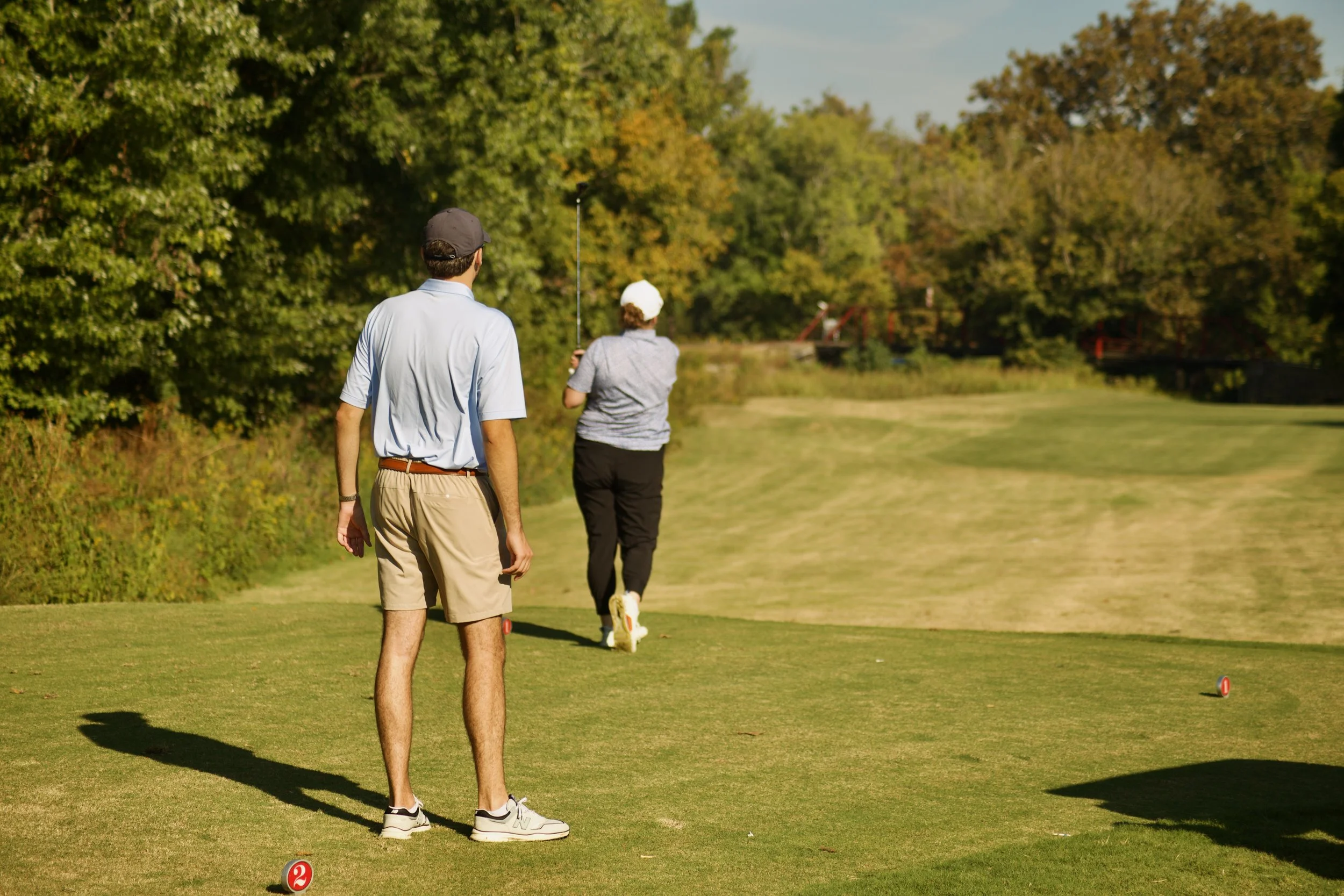 Two men playing golf on a lush green golf course, one of them is swinging a golf club while the other watches, surrounded by trees under a clear sky.