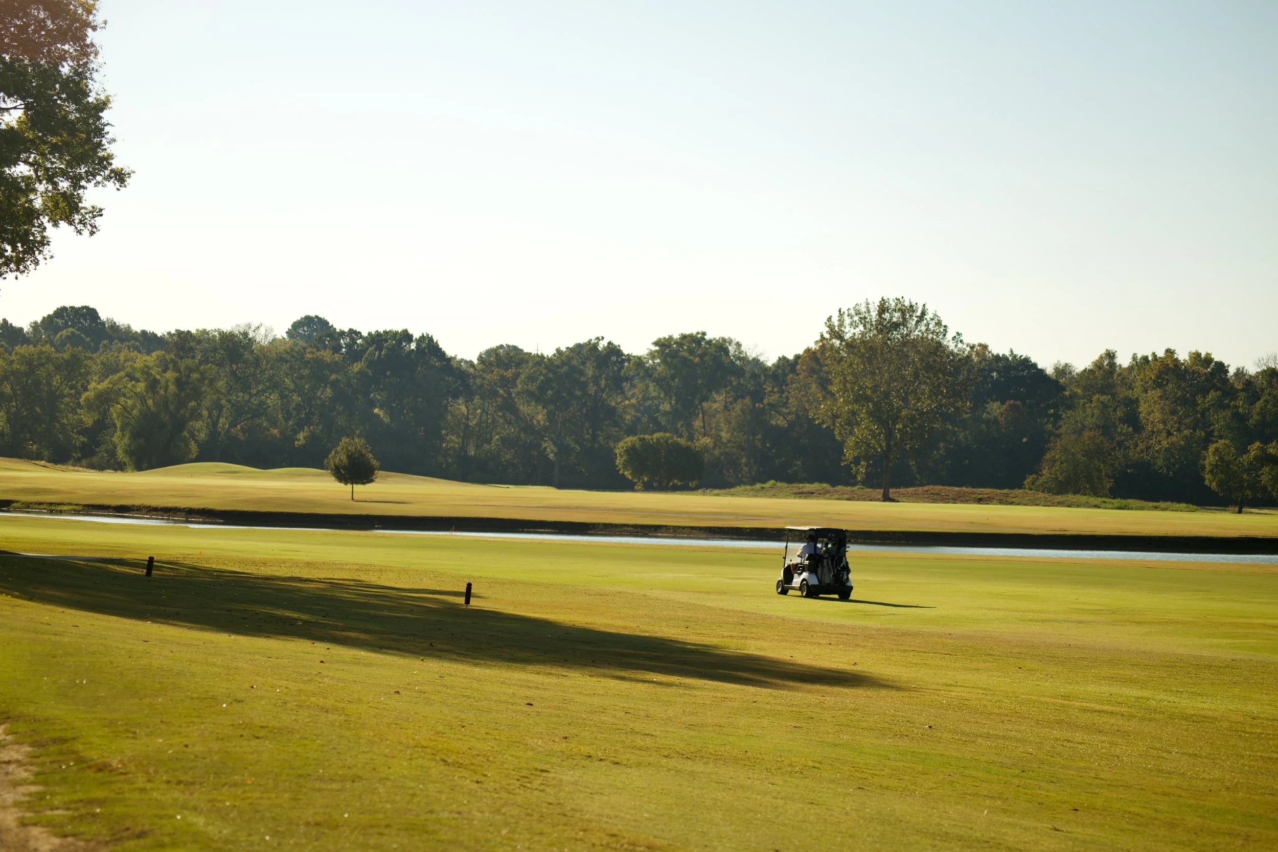 A golf course with a cart parked on the grass near water, surrounded by trees under a clear sky.