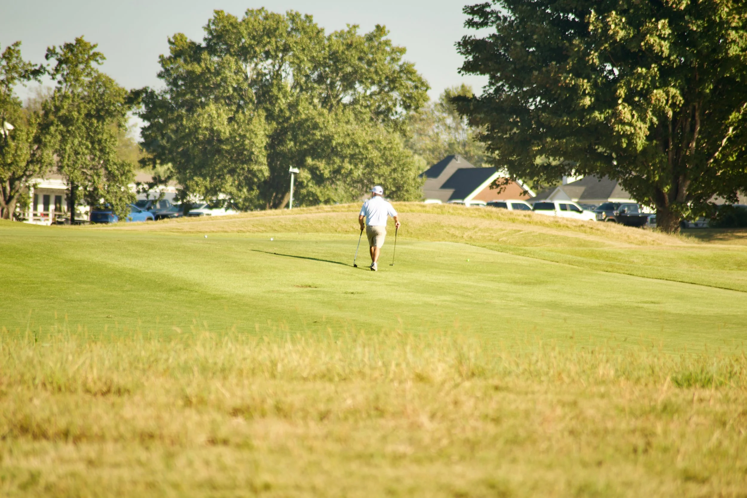 A person walking on a golf course with golf clubs in hand, surrounded by trees and houses in the background.