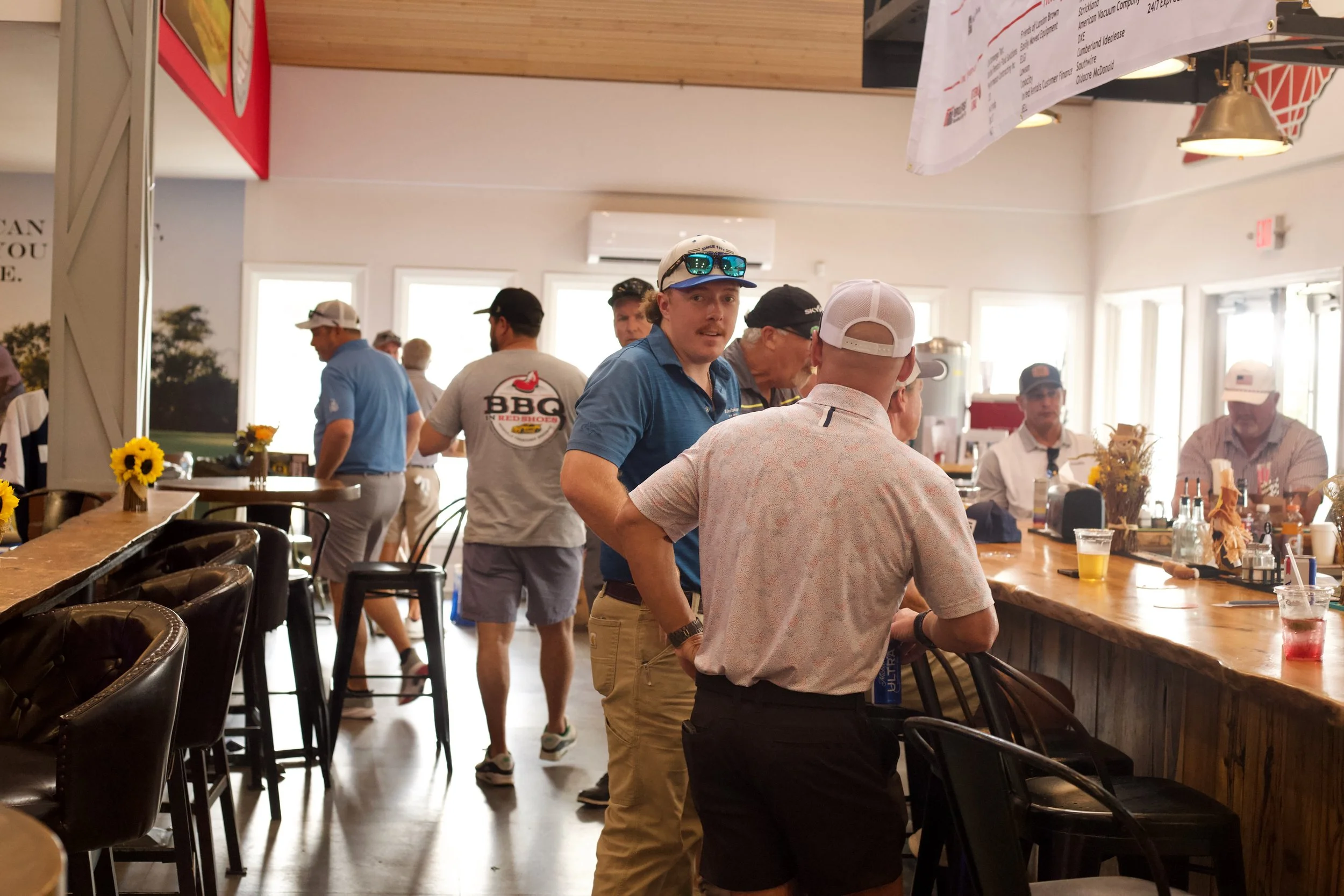 People gathering inside a restaurant or bar with a wooden ceiling, large windows, sunflower decorations, and a bar area with chairs. Some are standing and chatting while others are seated at the counter.