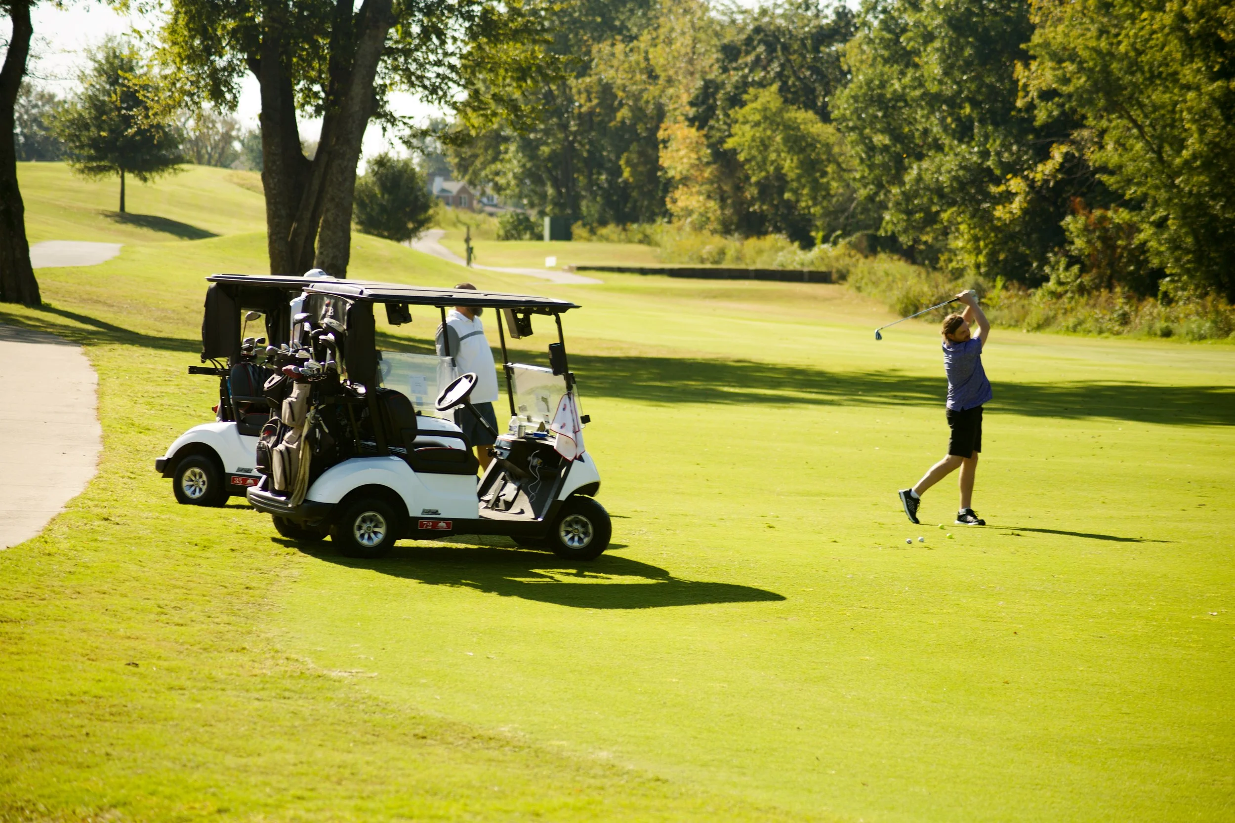 A person playing golf on a sunny day, with a golf cart carrying golf clubs nearby, on a lush green golf course surrounded by trees.