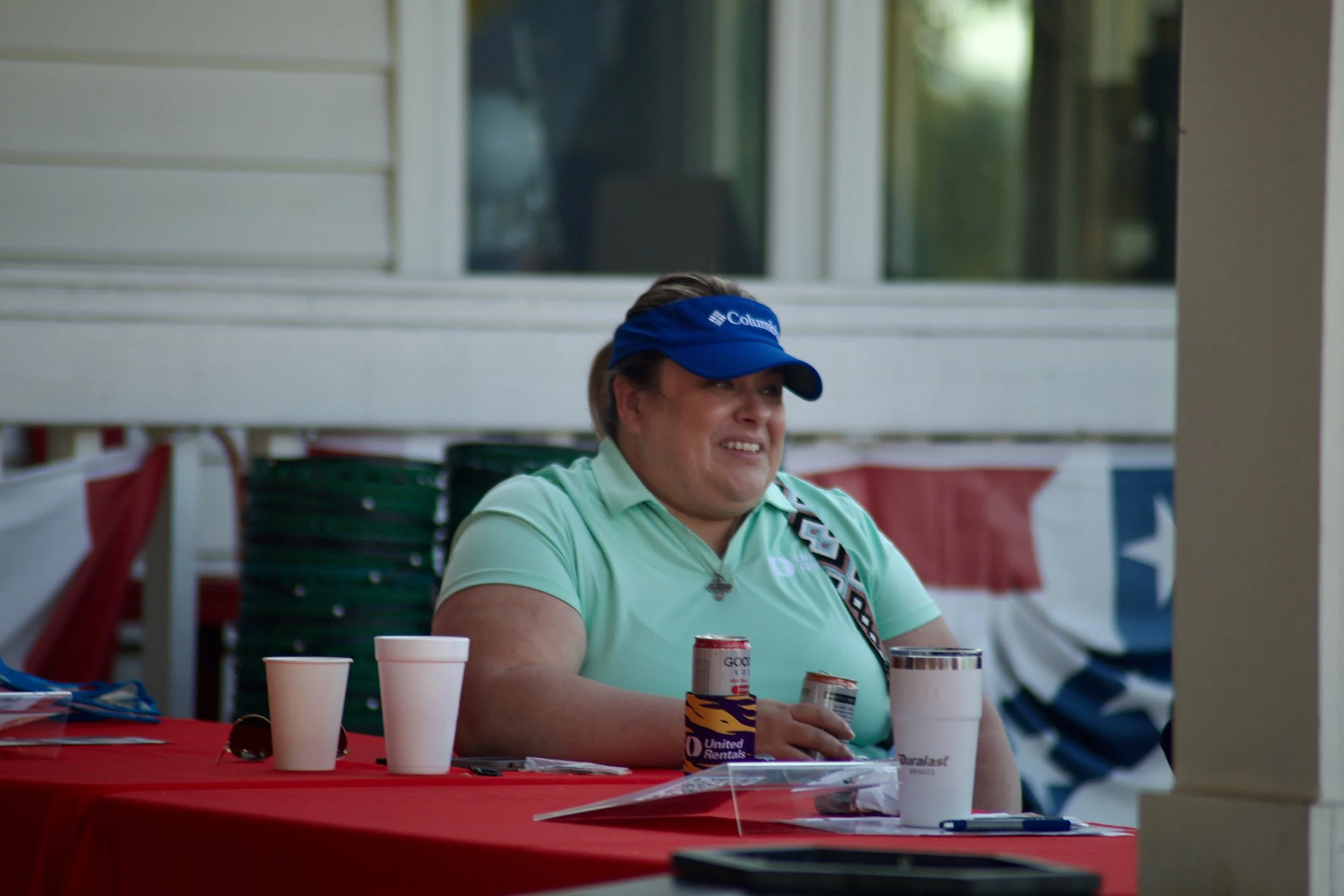 A woman in a cycling cap sitting at a table with cups and cans, smiling and outdoors.