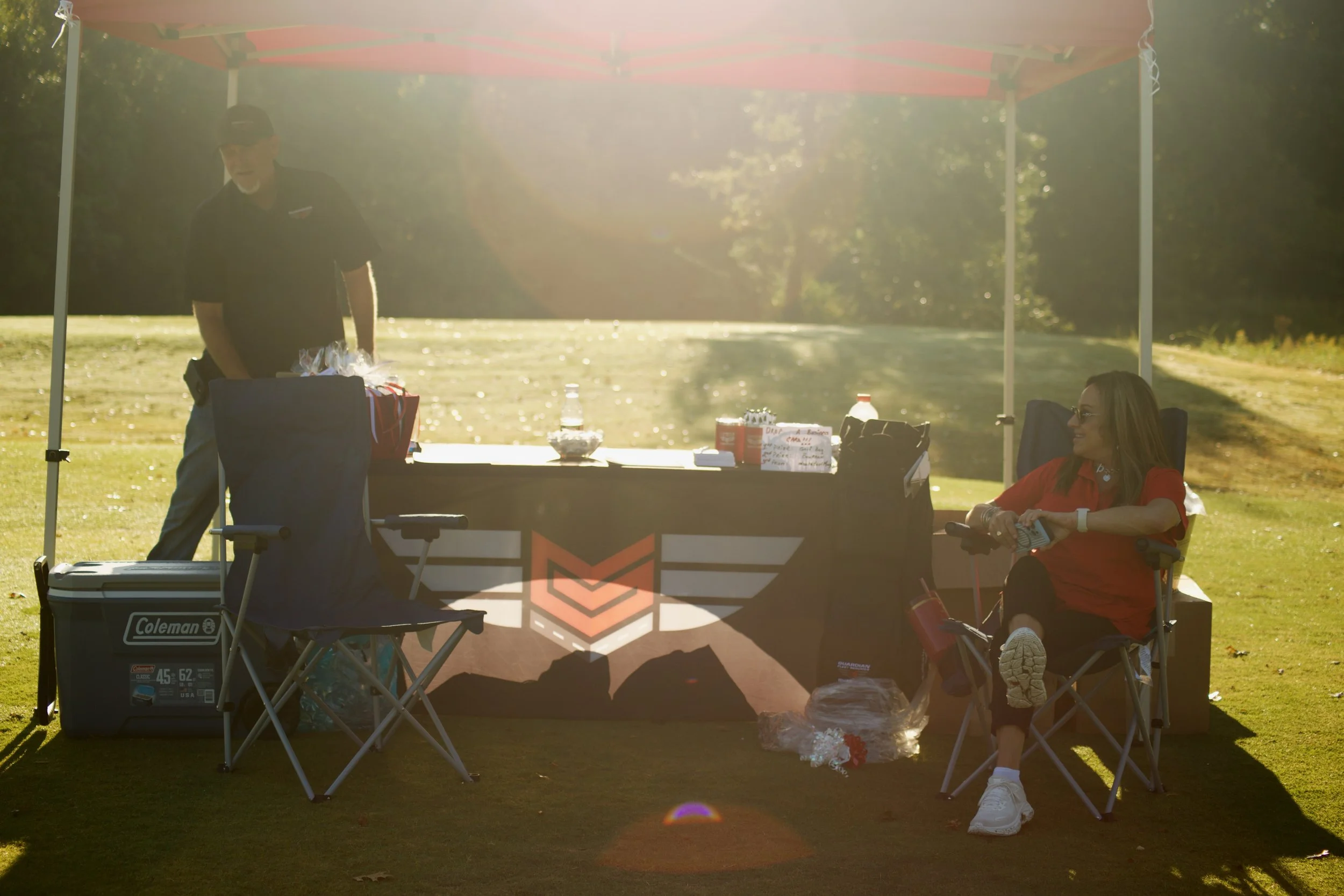 A man in a black shirt and cap and a woman in a red shirt and glasses sitting in chairs under a red canopy outdoors on a sunny day. There is a table with snacks and drinks between them, and the background features a grassy area and trees.