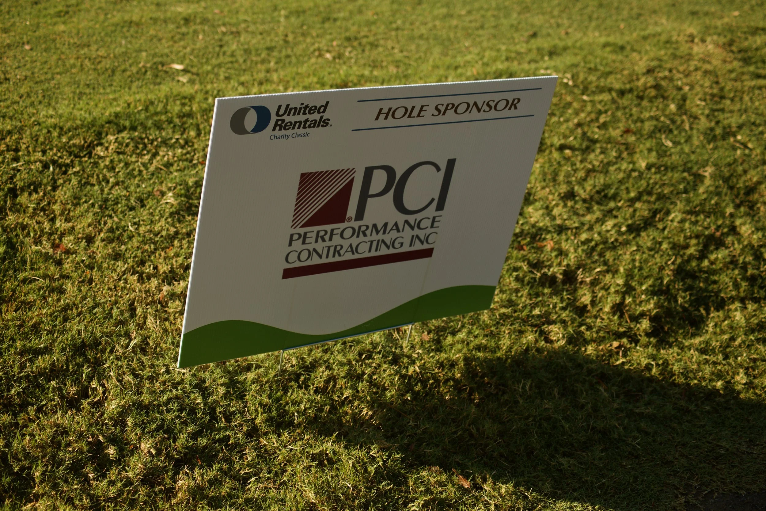 A sign on a golf course with sponsorship information and logos, positioned on a grassy area.