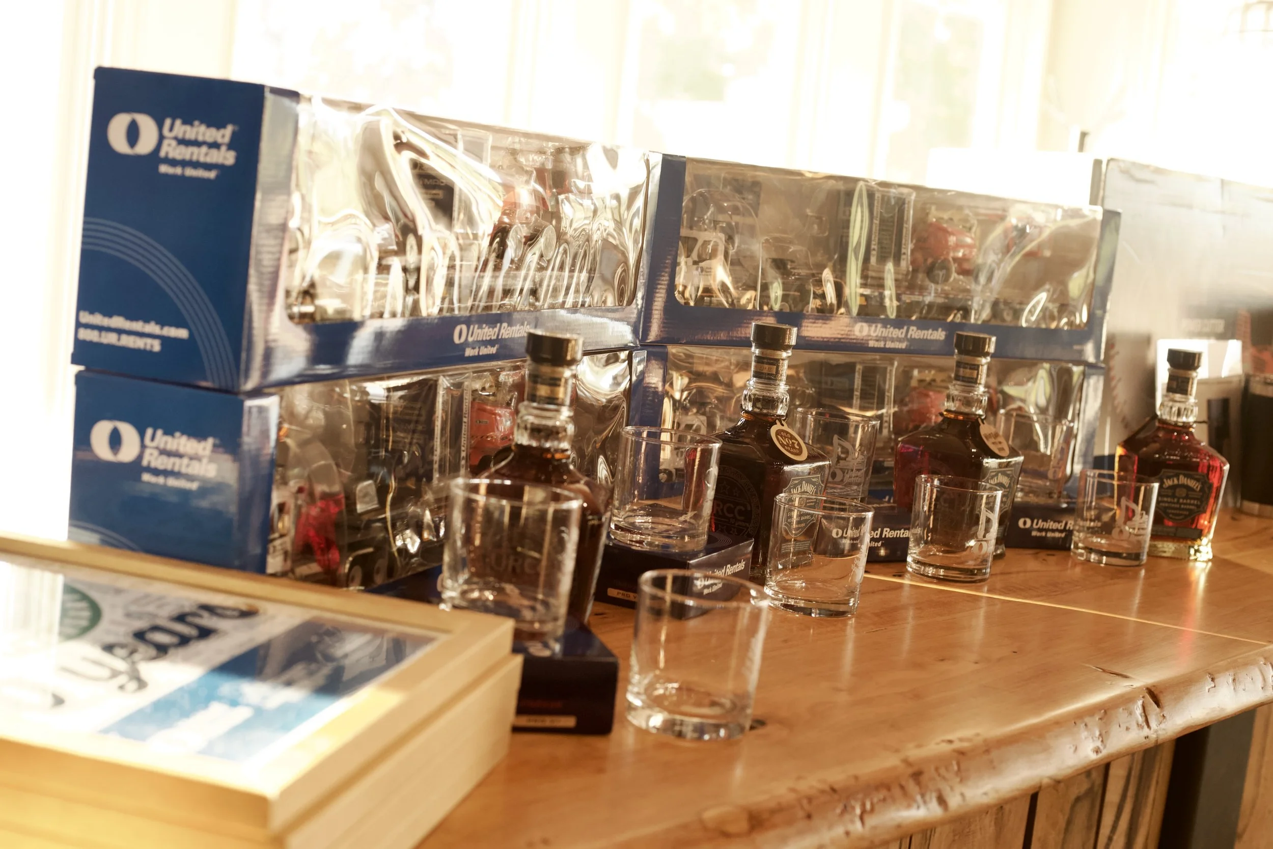 Stacked boxes of United Rentals with clear glass bottles and shot glasses on a wooden table in a well-lit room.