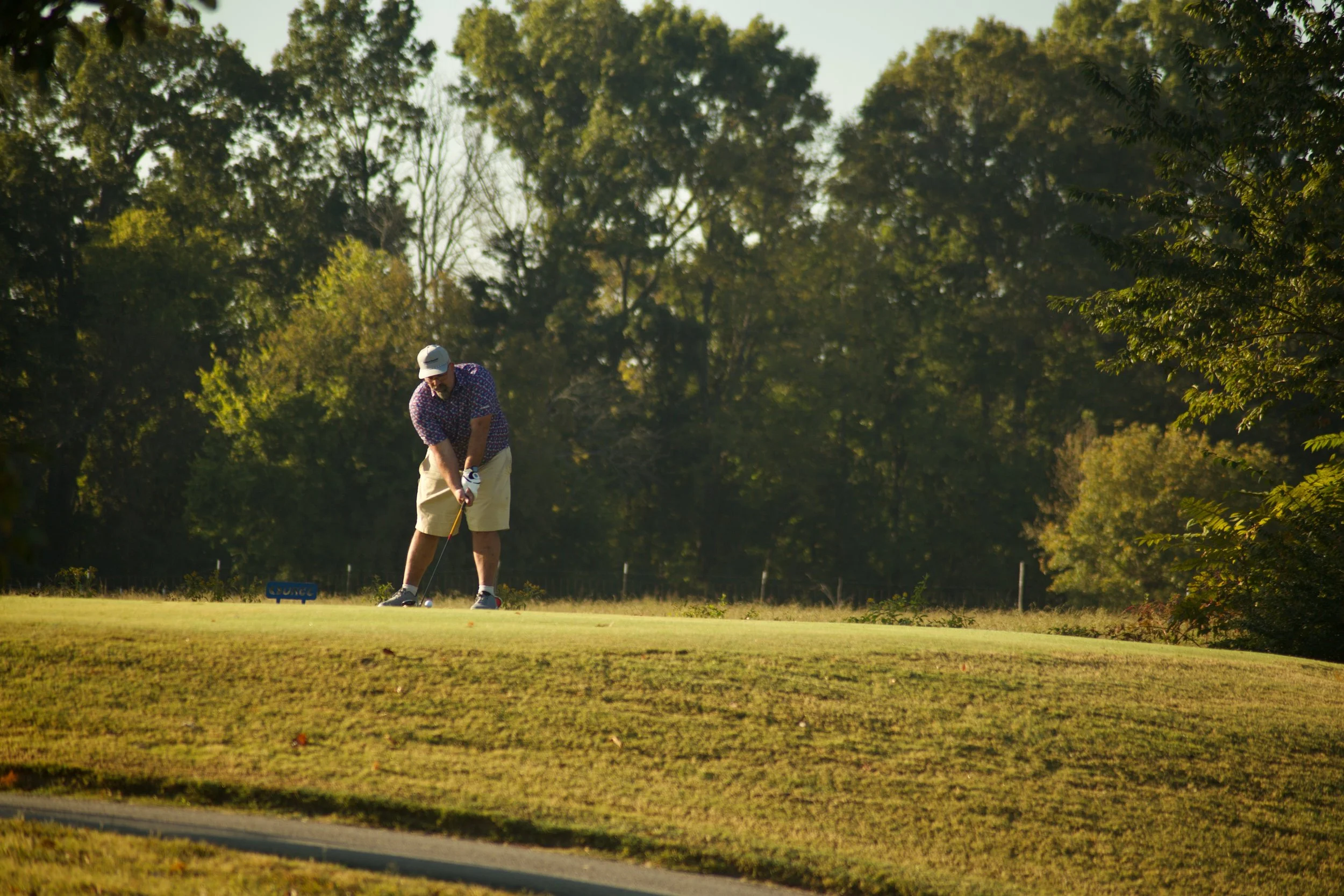 A man in a hat and patterned shirt preparing to hit a golf ball on a golf course surrounded by trees during daylight.