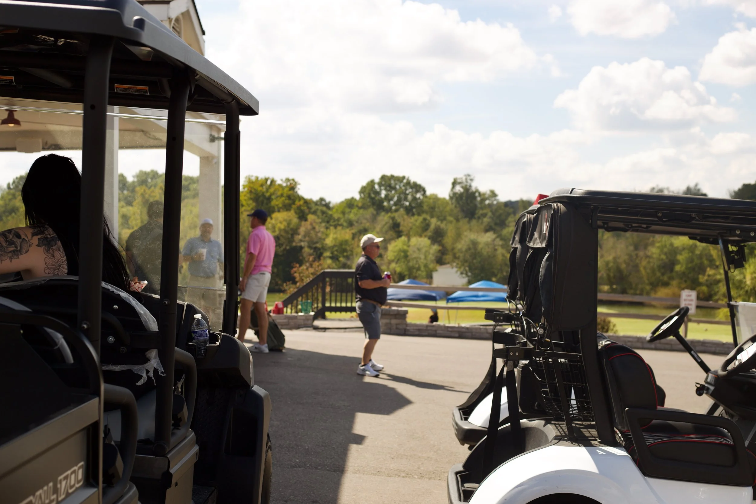 Golf carts parked at an outdoor setting with people standing and walking in the background, trees and a partly cloudy sky visible.