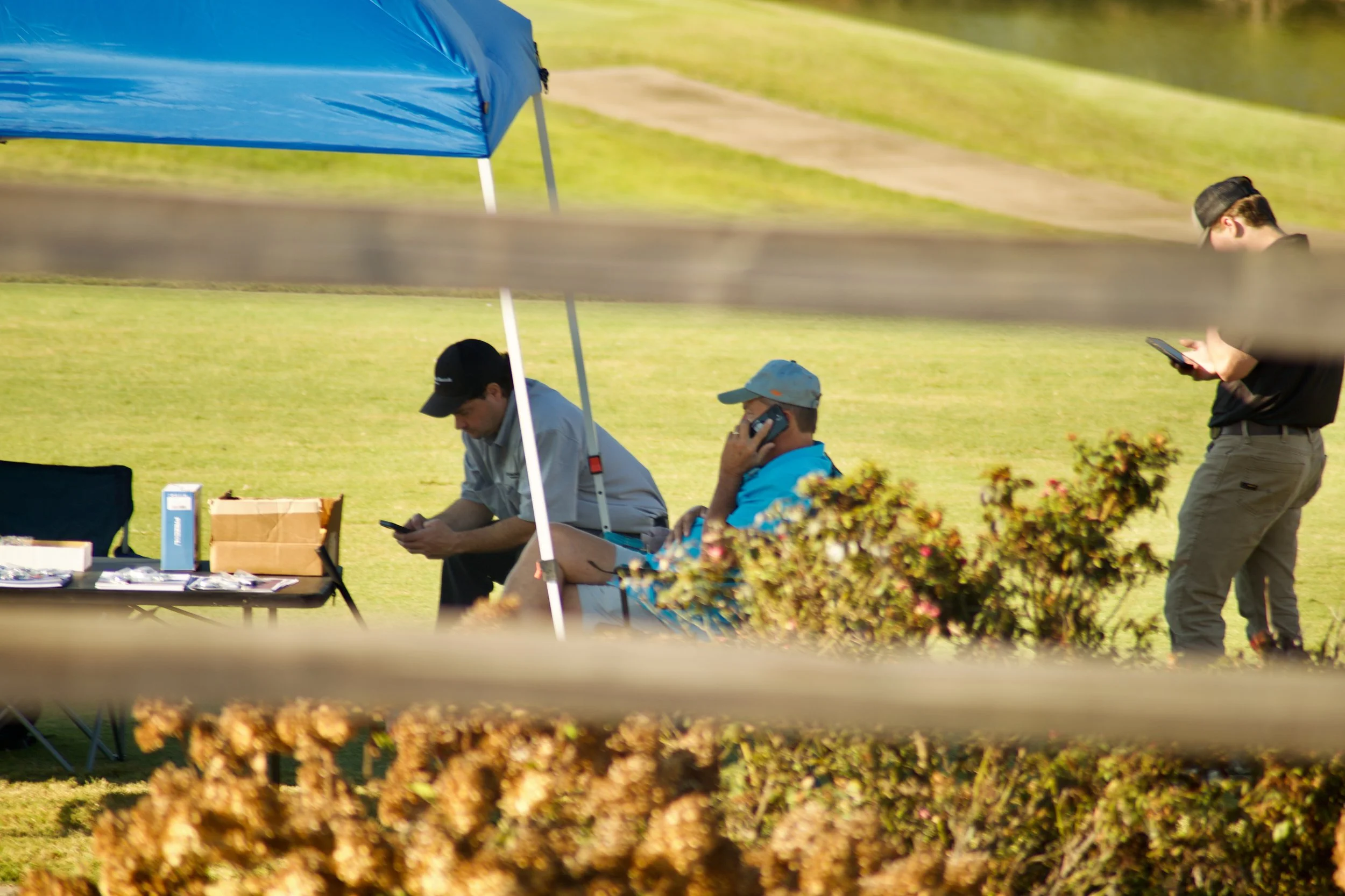 People sitting and standing under a blue canopy tent outdoors on a grassy area, with two individuals using phones and a table with items.