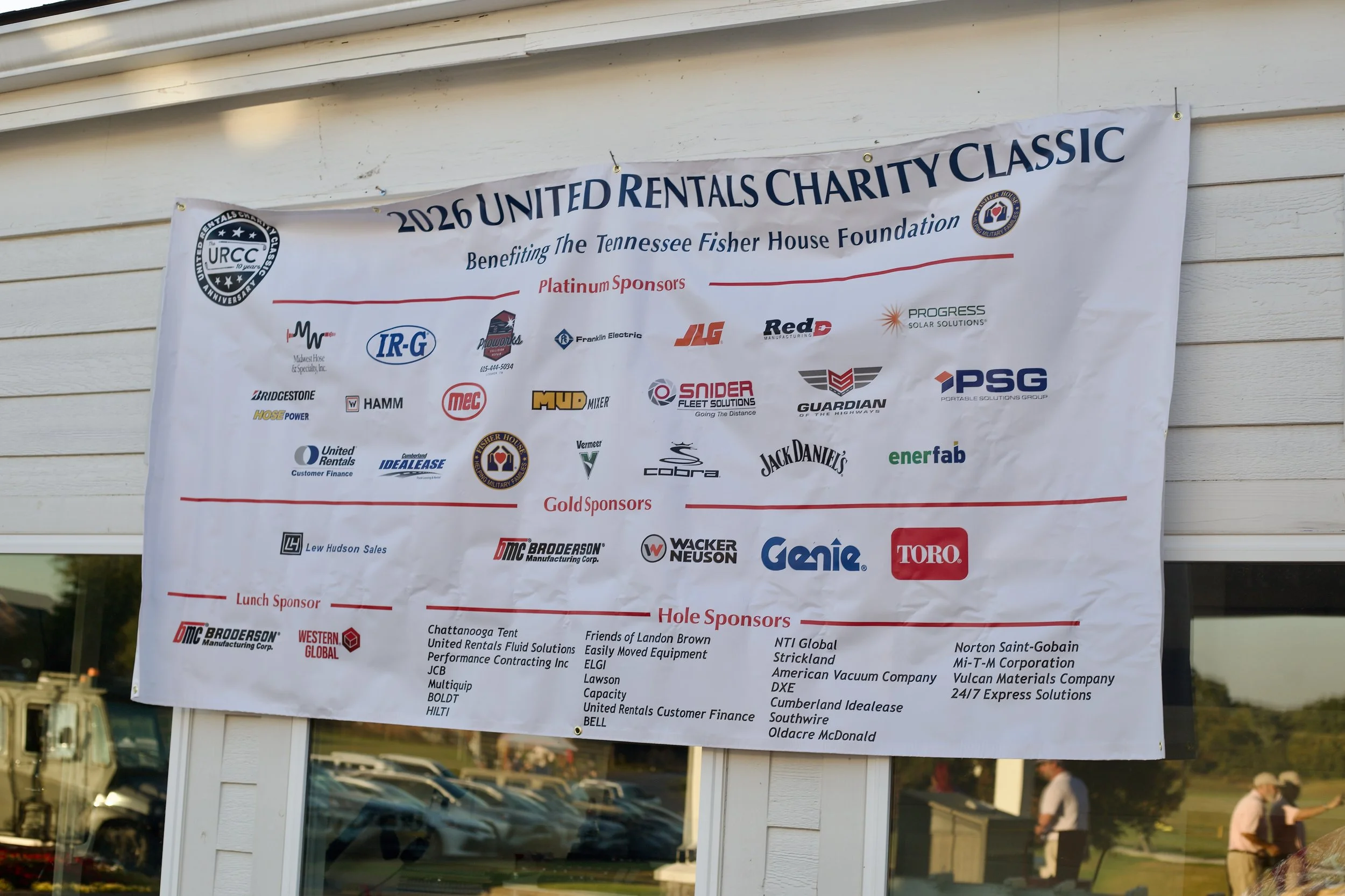 Banner for the 2026 United Rentals Charity Classic, benefiting the Tennessee Fisher House Foundation, displayed outside a building with people and parked cars visible in the reflection of the glass.
