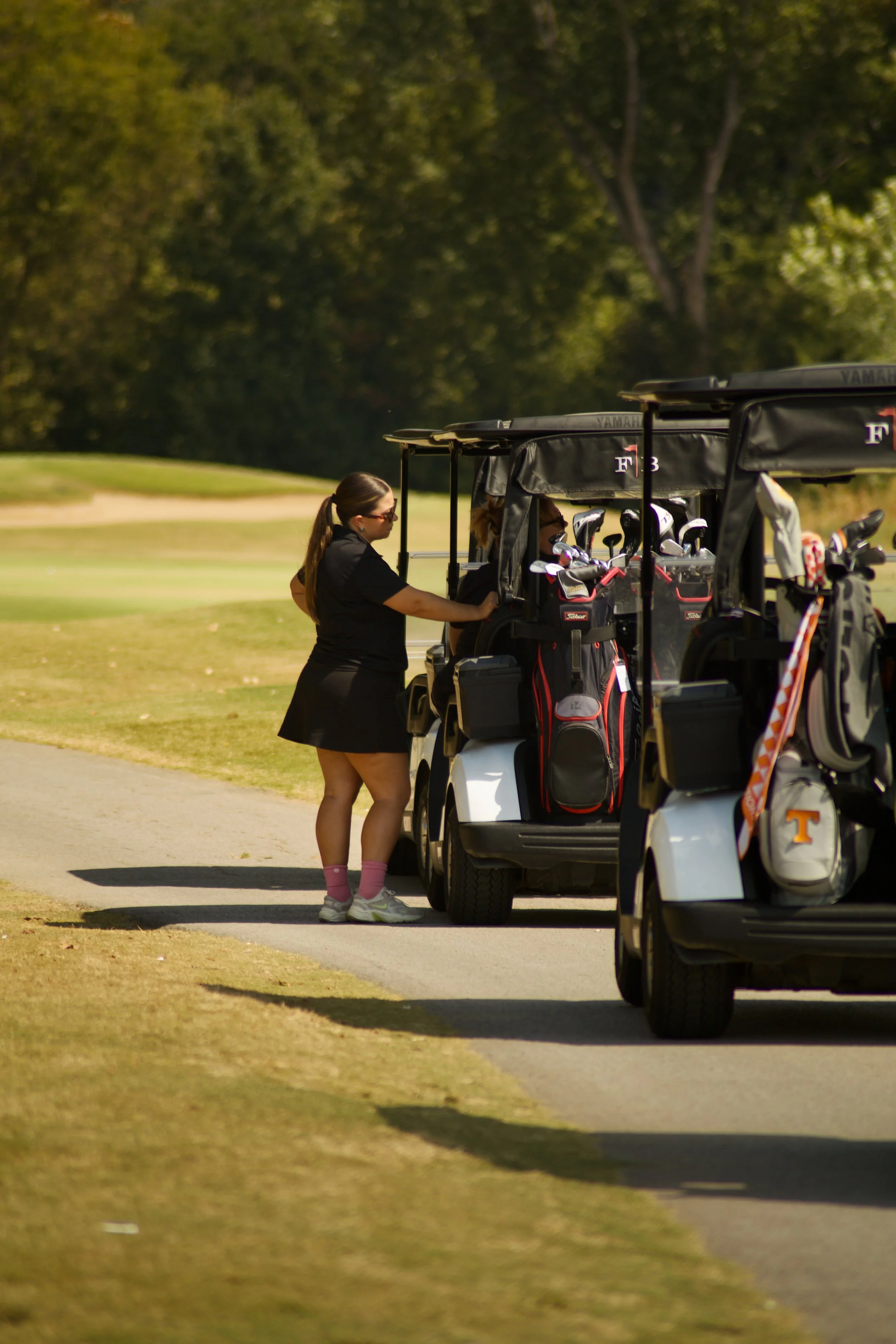 A woman in black attire and pink socks is putting clubs into a golf cart filled with golf clubs and bags, on a golf course with trees in the background.