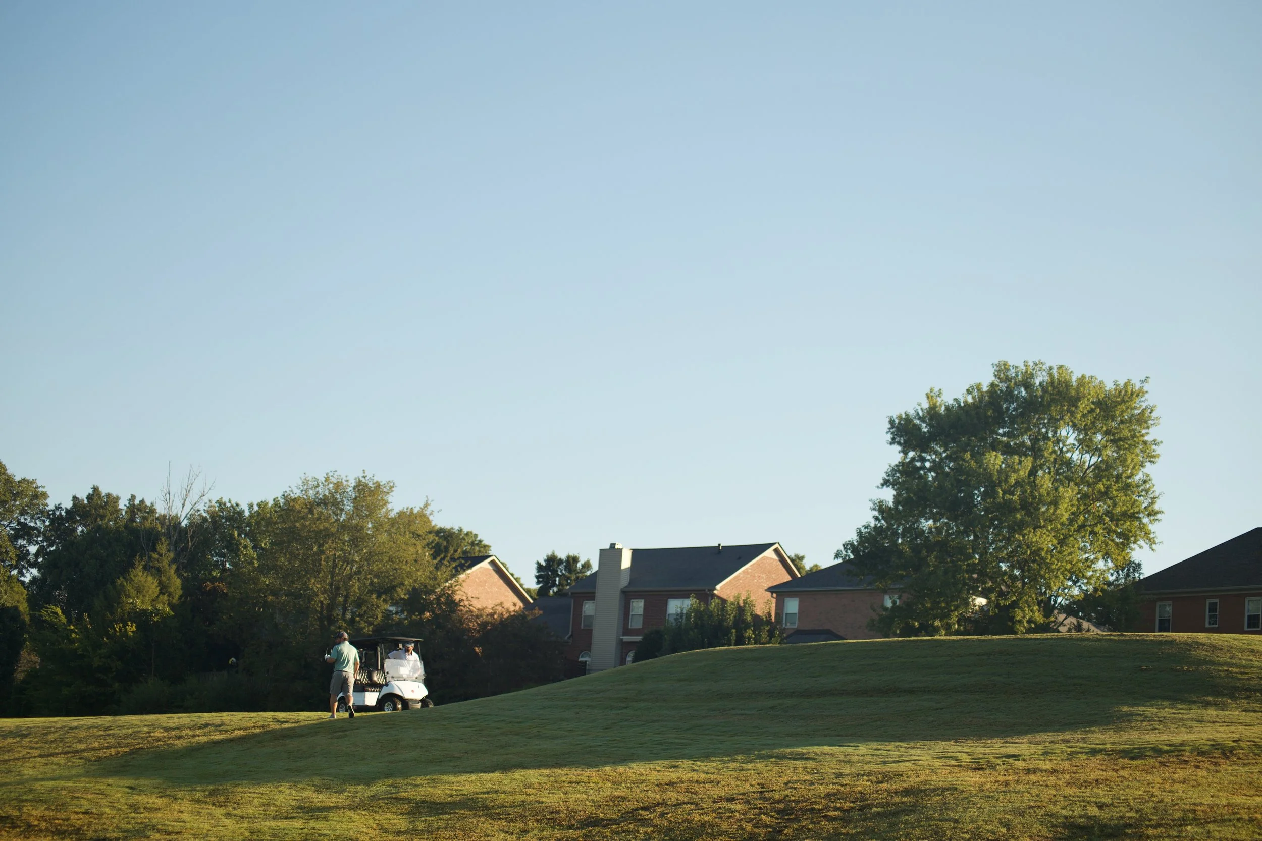 A man walking on a golf course near a golf cart, with houses and trees in the background under a clear blue sky.