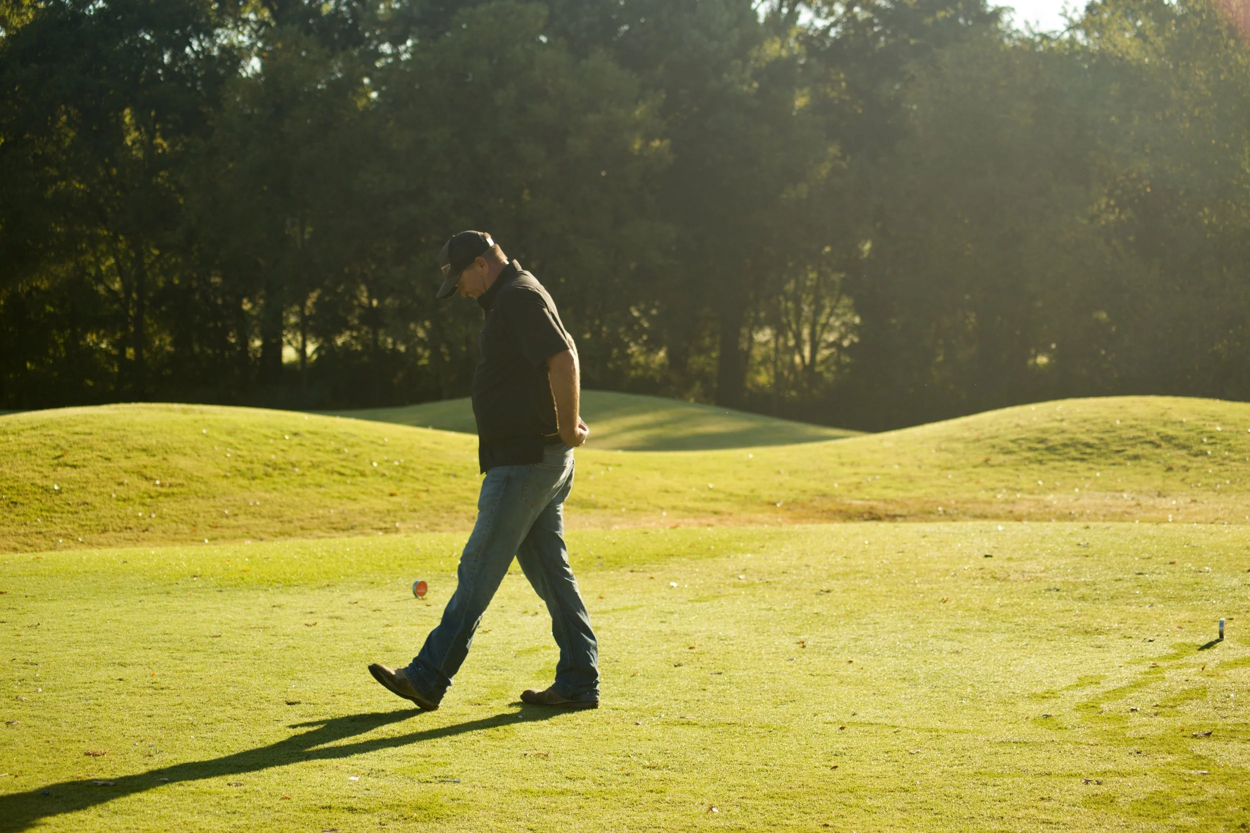A man wearing a black cap, black shirt, and jeans walking on a golf course with trees in the background during daytime.