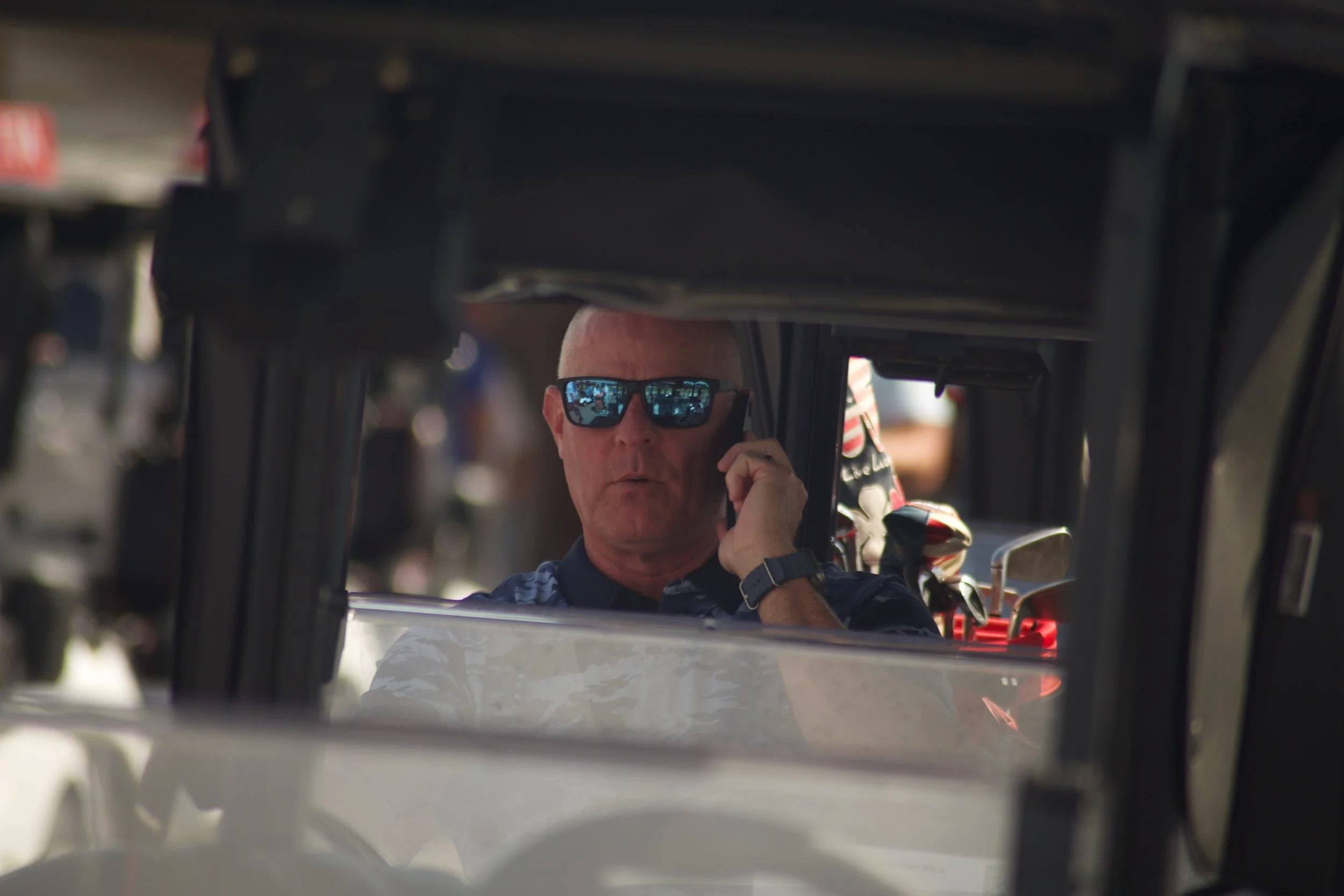 A man with sunglasses talking on a cellphone is seen through the window of a race car or similar vehicle.
